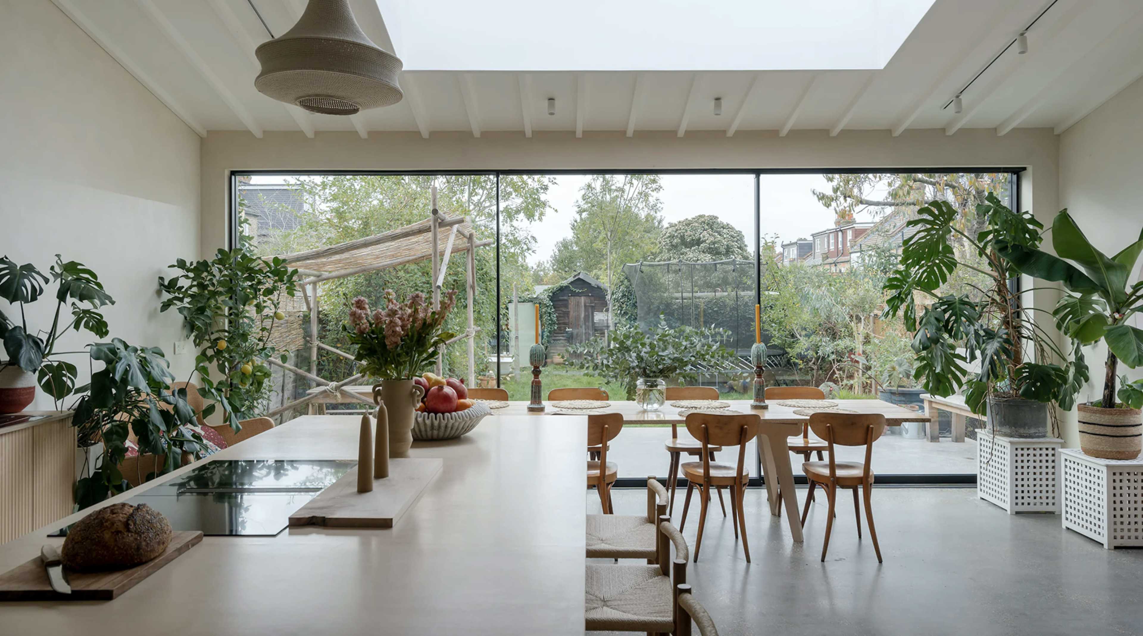 A modern kitchen with a long countertop overlooks a garden through large glass windows, featuring various indoor plants and wooden dining chairs.
