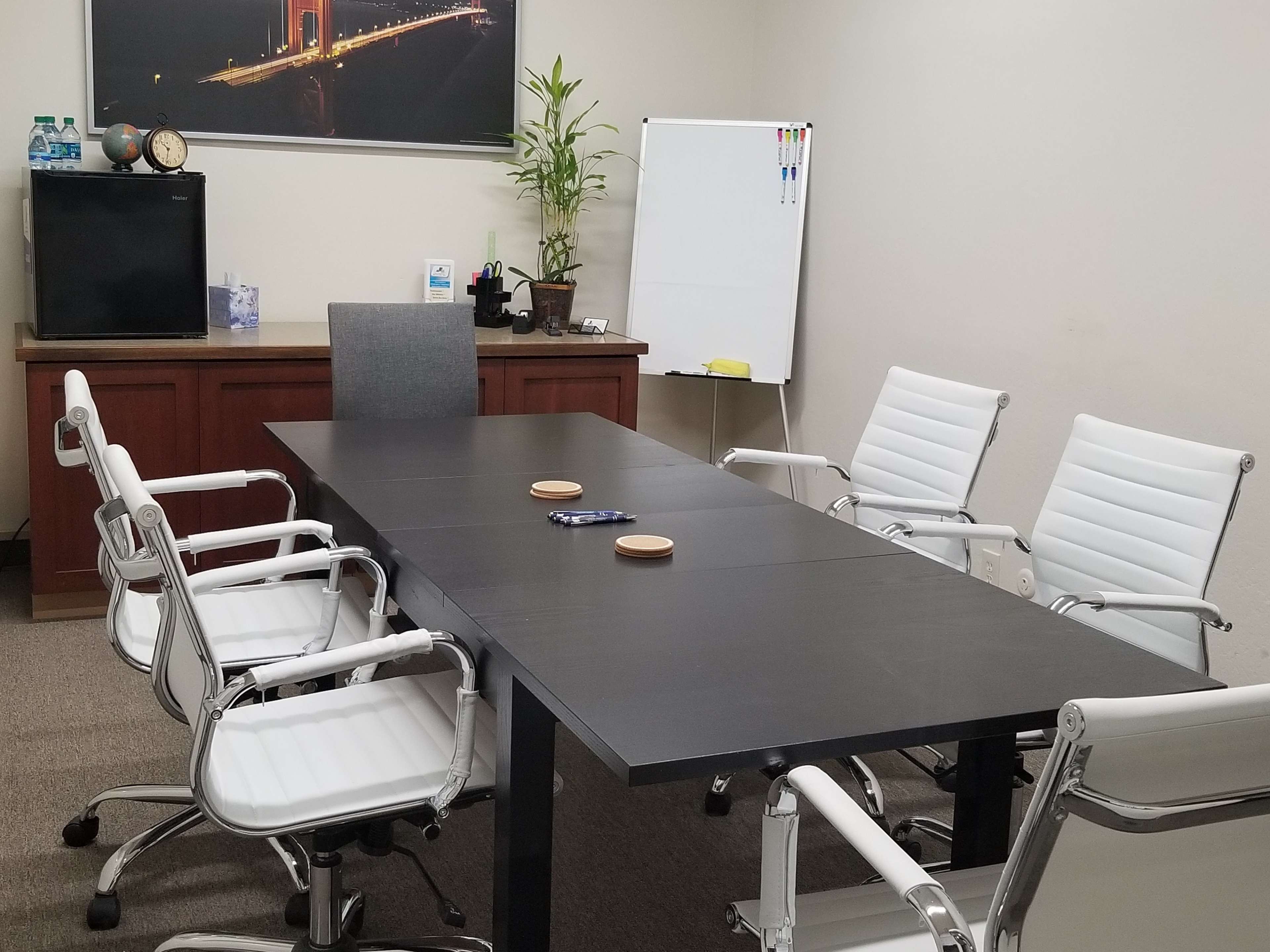 A modern conference room features a black table surrounded by white chairs, with a wall-mounted picture, a plant, and a whiteboard in the background.