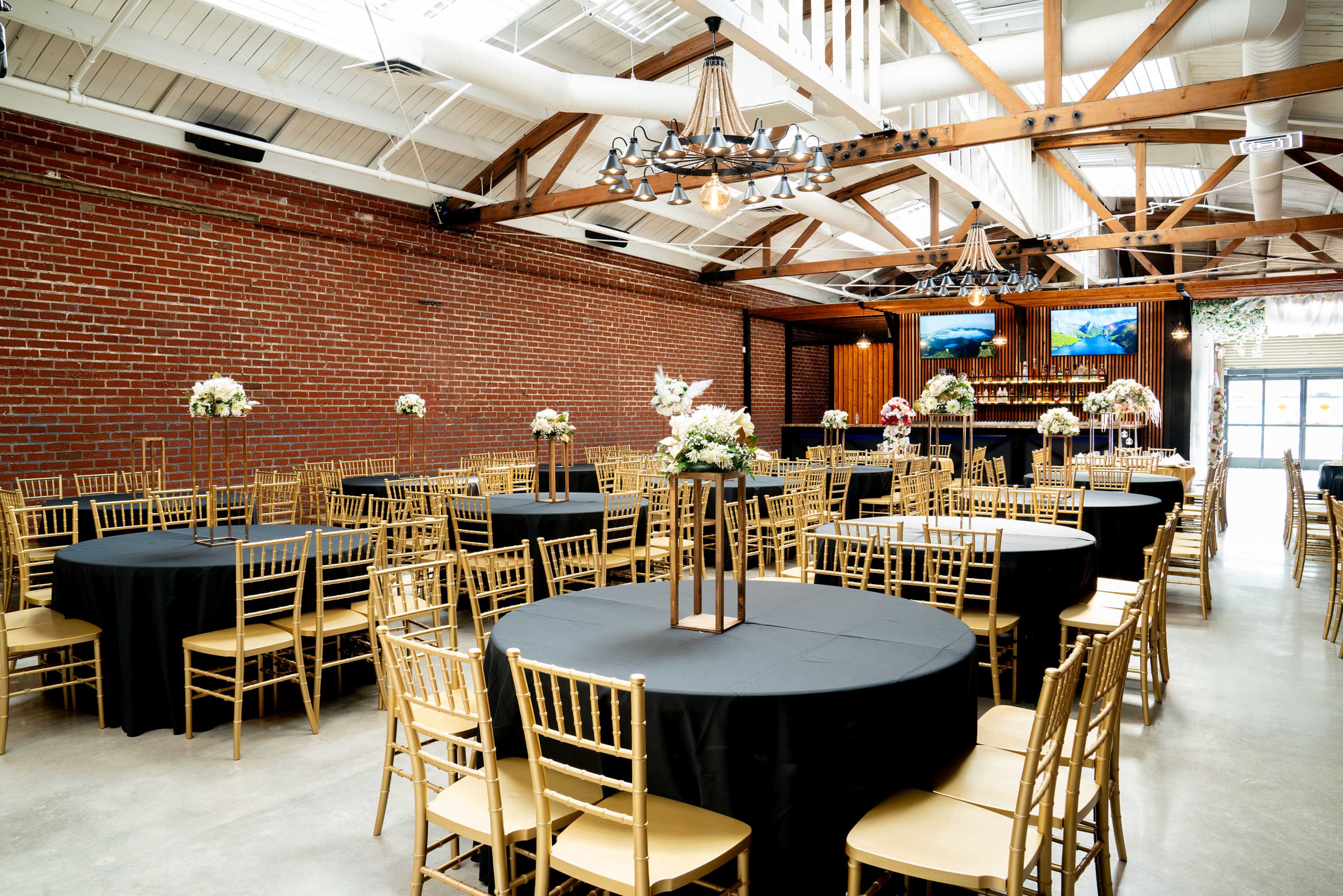 The image shows a spacious event venue with round tables covered in black linens and gold chairs, set against a backdrop of exposed red brick walls and wooden beams.