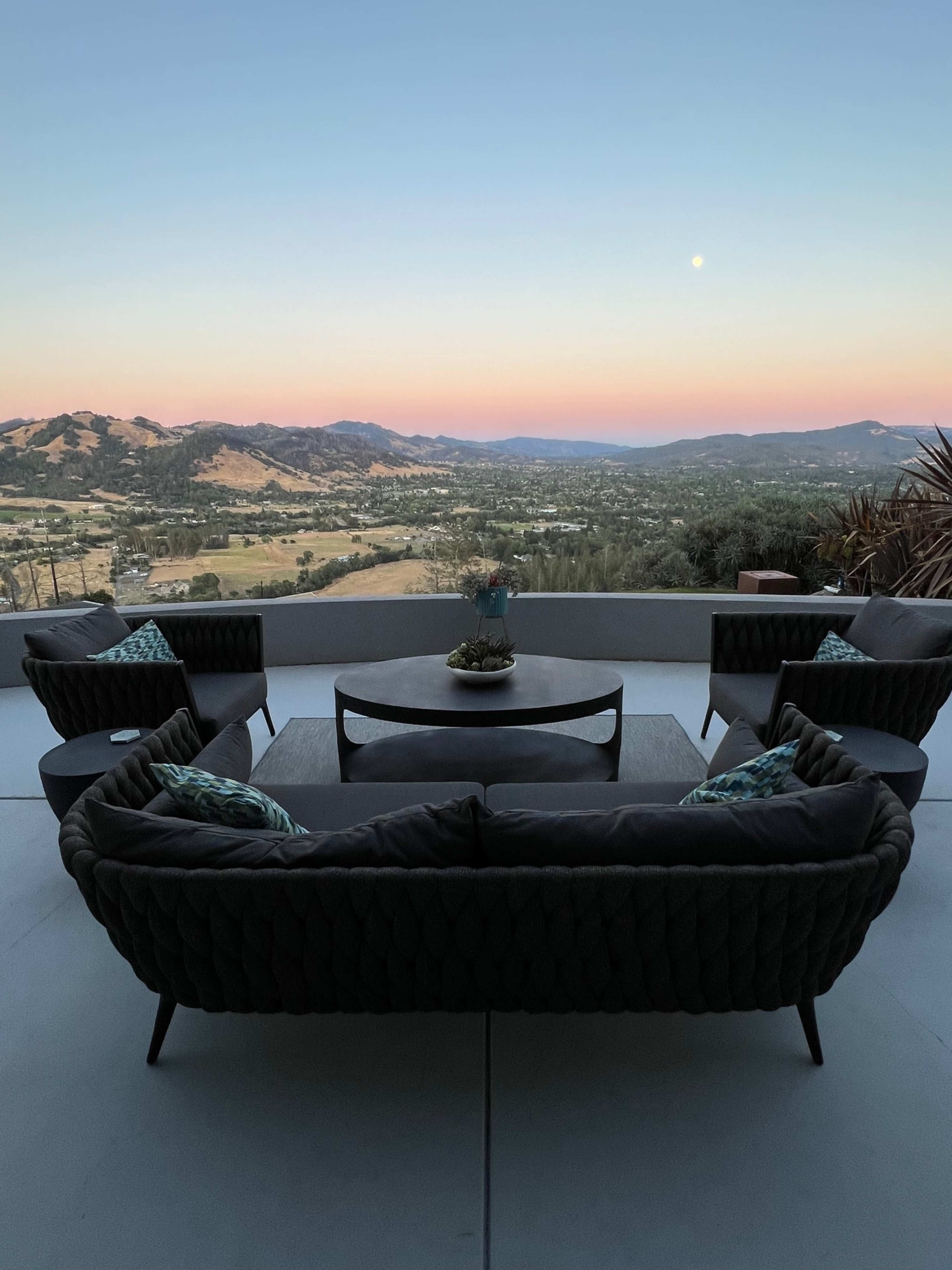 A seating area with four chairs and a round table overlooks a valley at dusk, with mountains in the background and a pale moon in the sky.