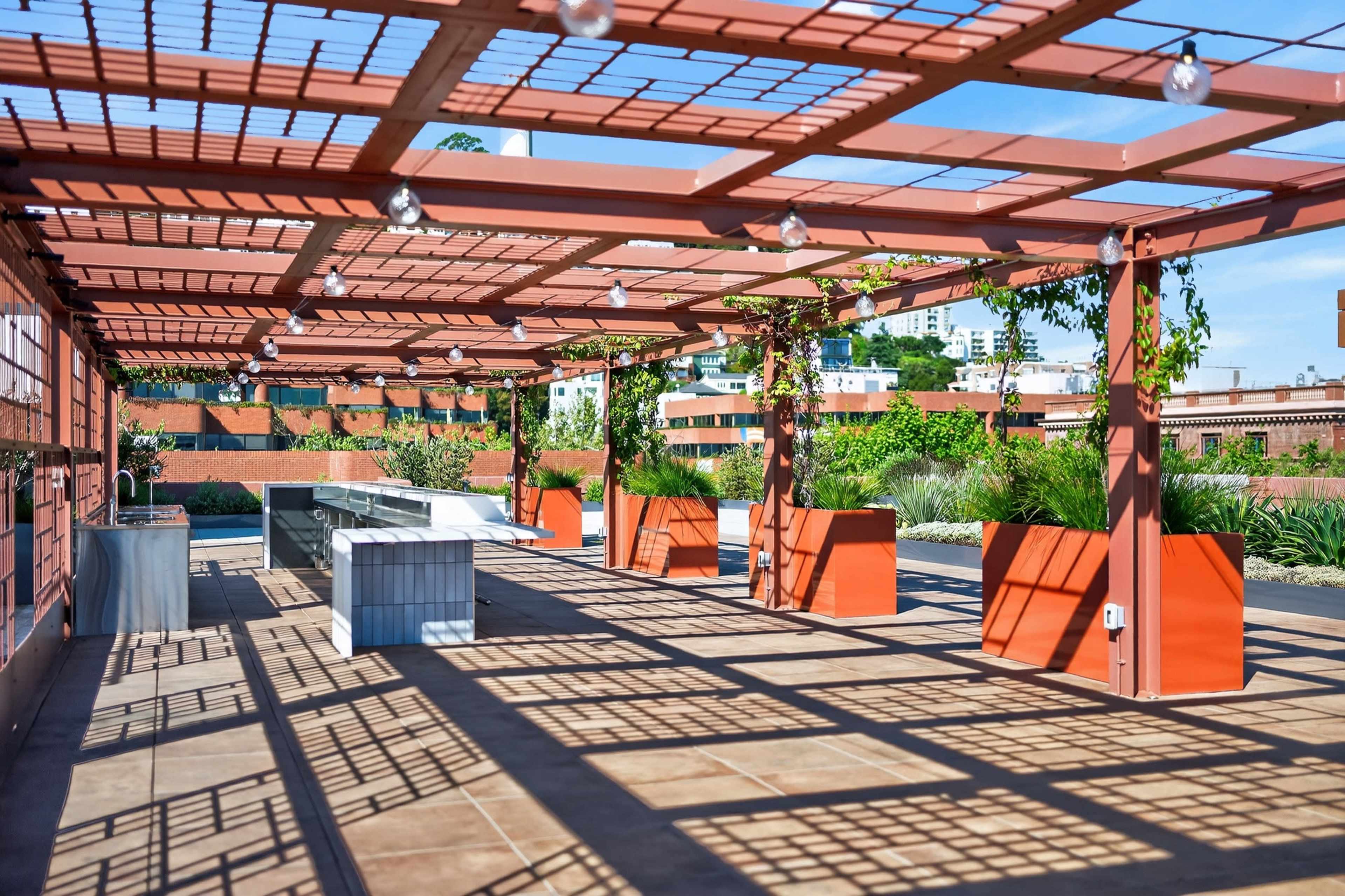 The image shows a spacious outdoor rooftop area featuring wooden pergolas, seating arrangements, and decorative plants.