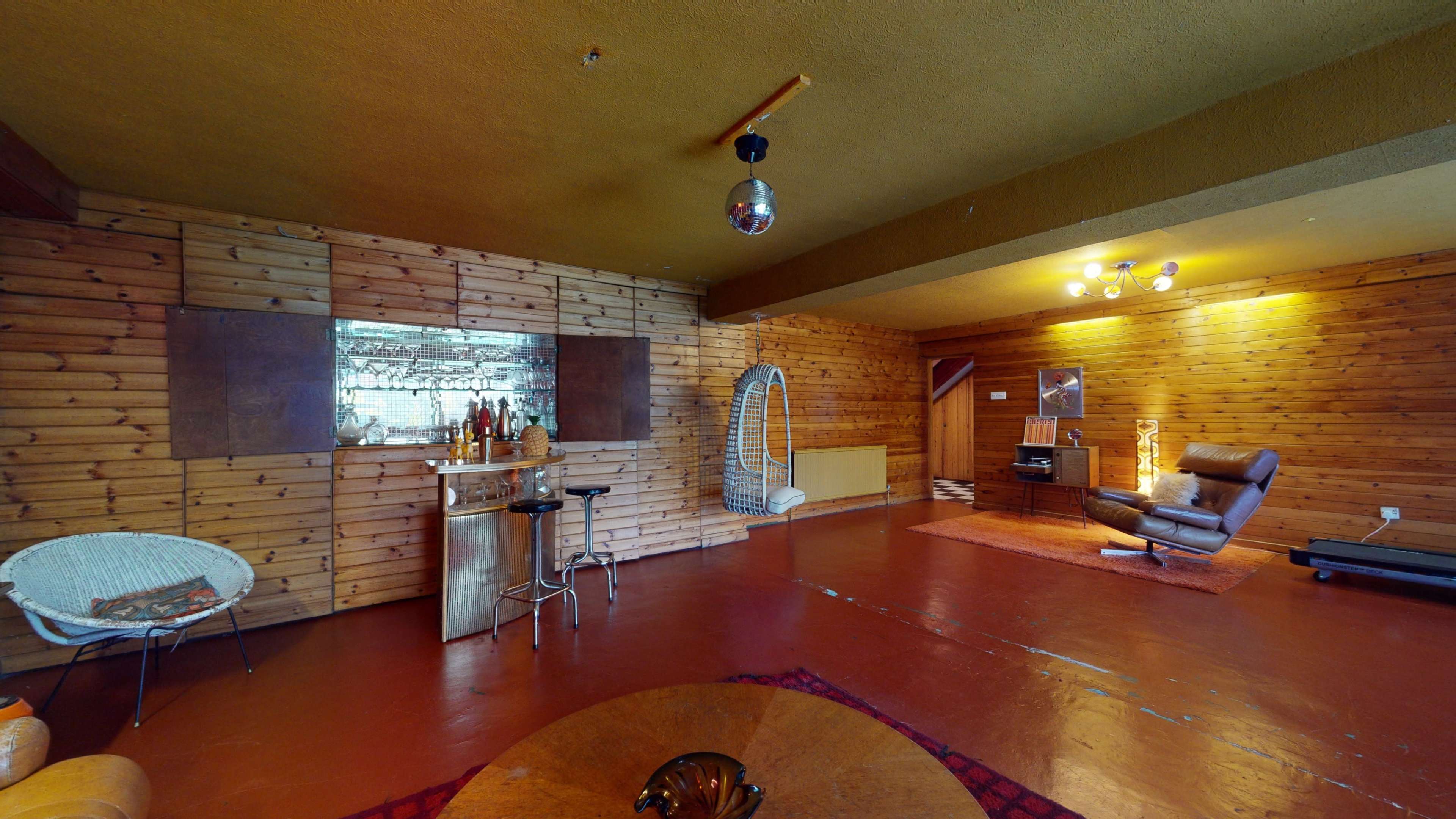 The image depicts a spacious interior featuring wooden walls, a bar area with drinks, a reclining chair, and various seating arrangements.