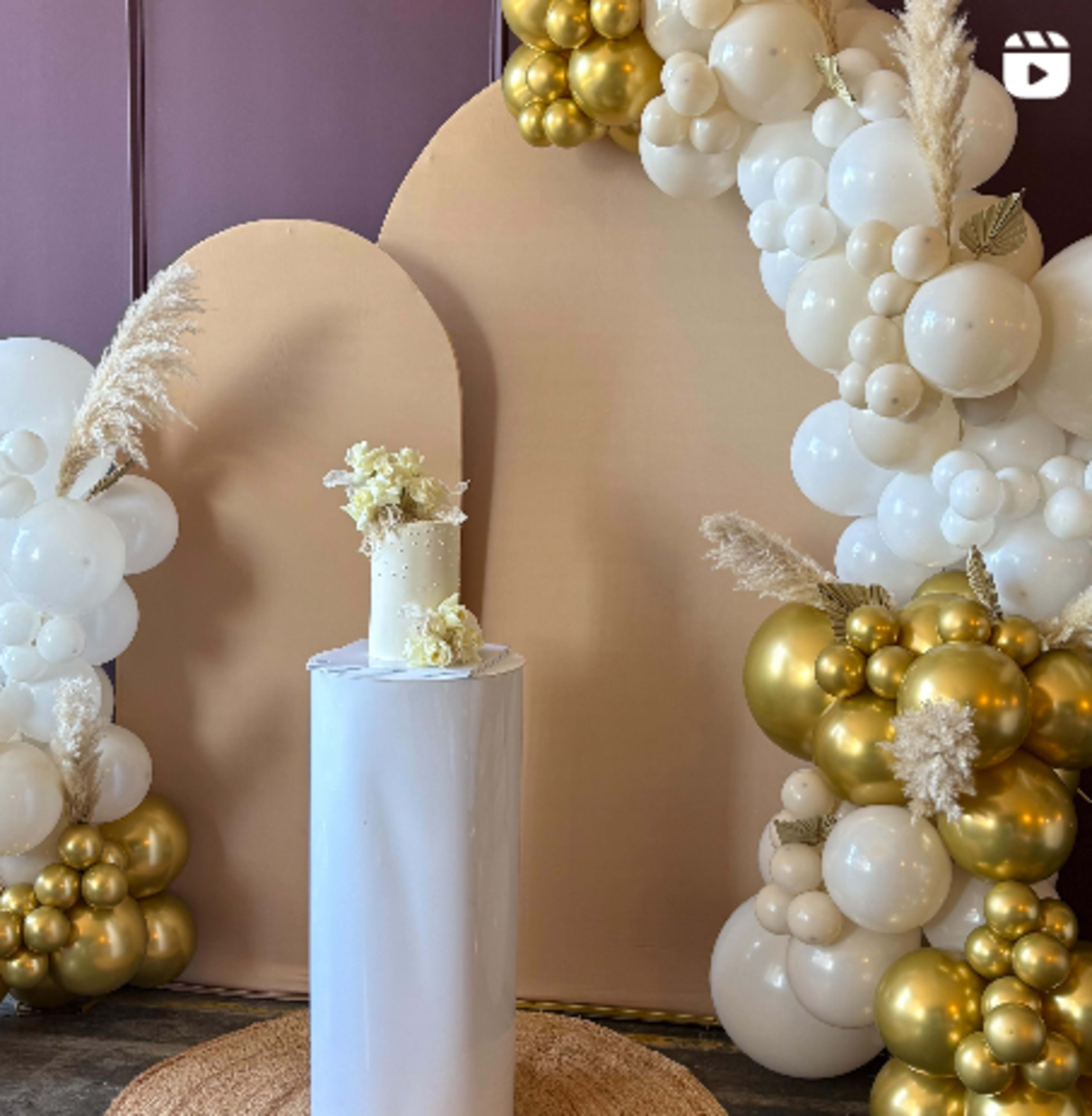 The image features a decorative backdrop made of balloons in white and gold, with a central white pedestal displaying a floral arrangement.