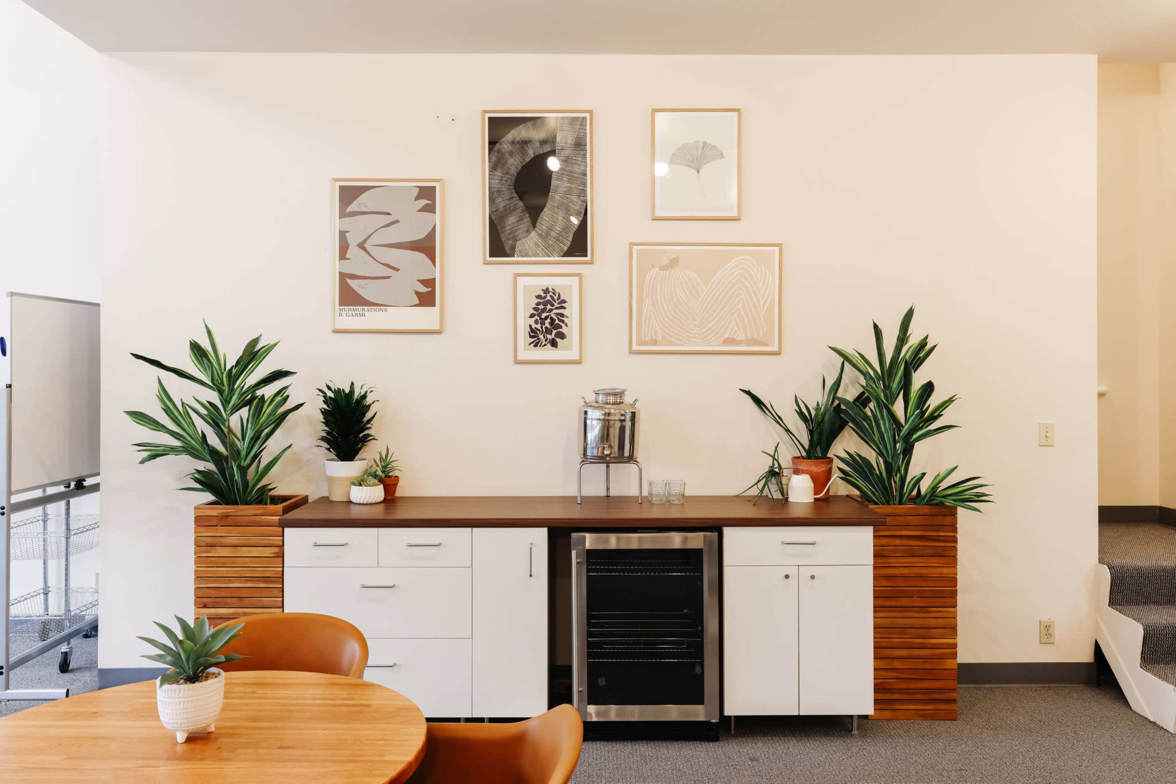 A modern kitchen area features a wooden countertop, a beverage cooler, and decorative plants alongside framed art on the wall.