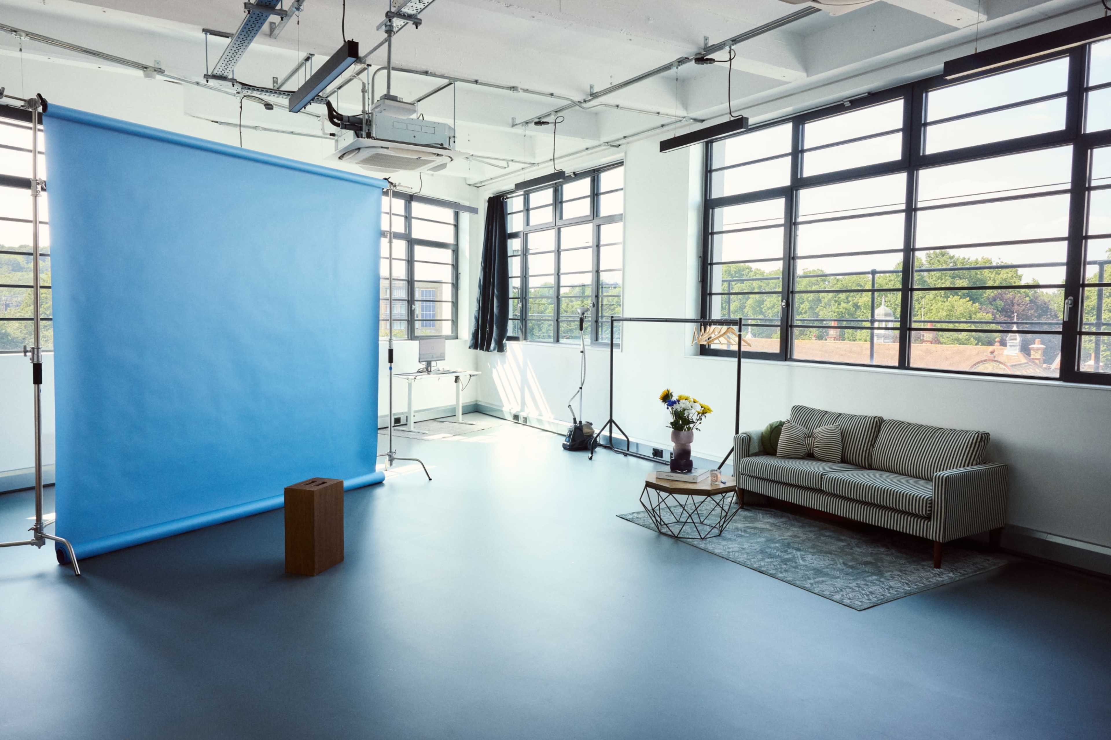 The image shows a bright studio space featuring a blue backdrop, a sofa, a coffee table, and large windows allowing natural light to enter.
