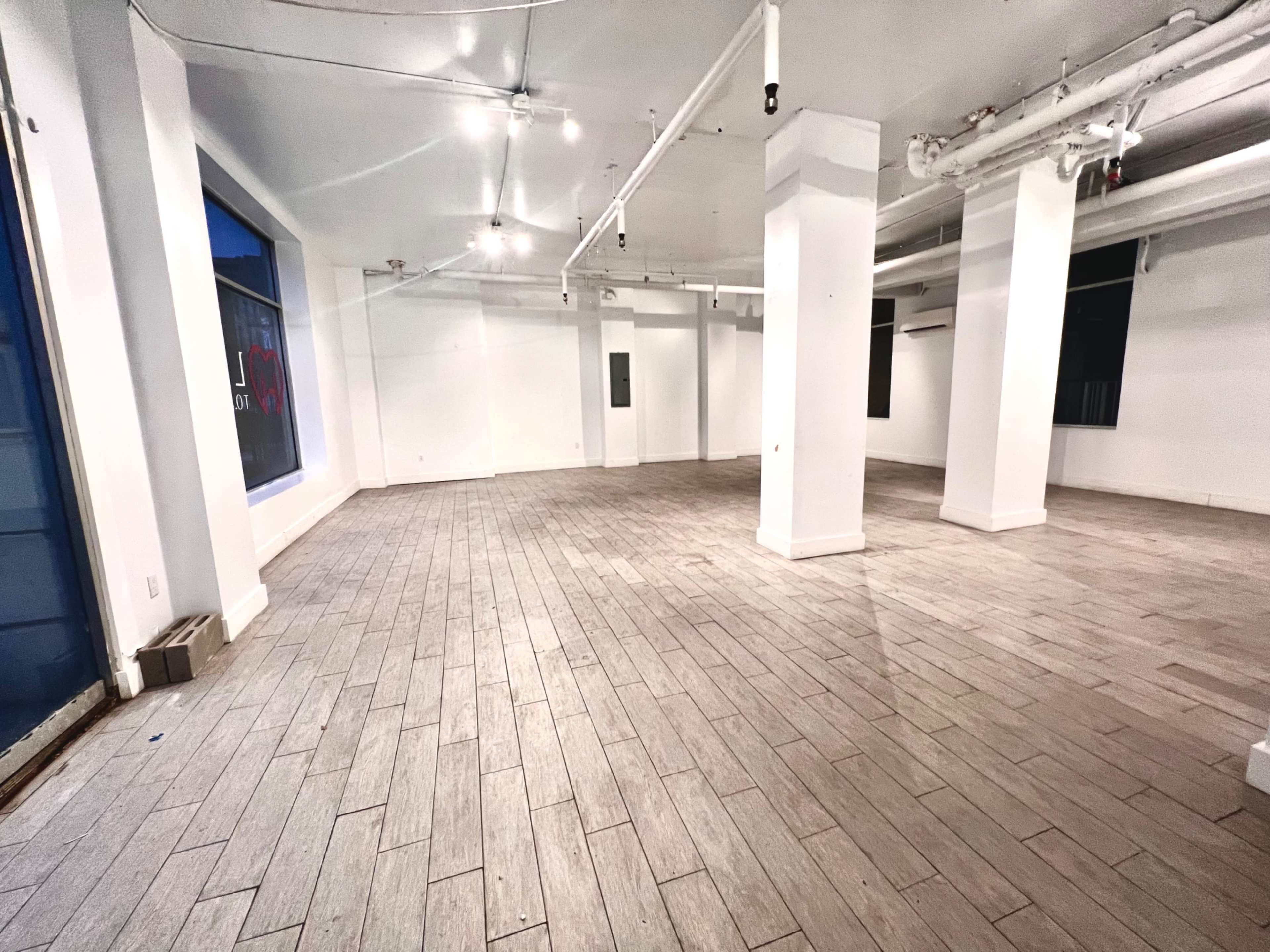 A spacious, empty commercial interior with white walls, gray tiled flooring, and several ceiling lights.