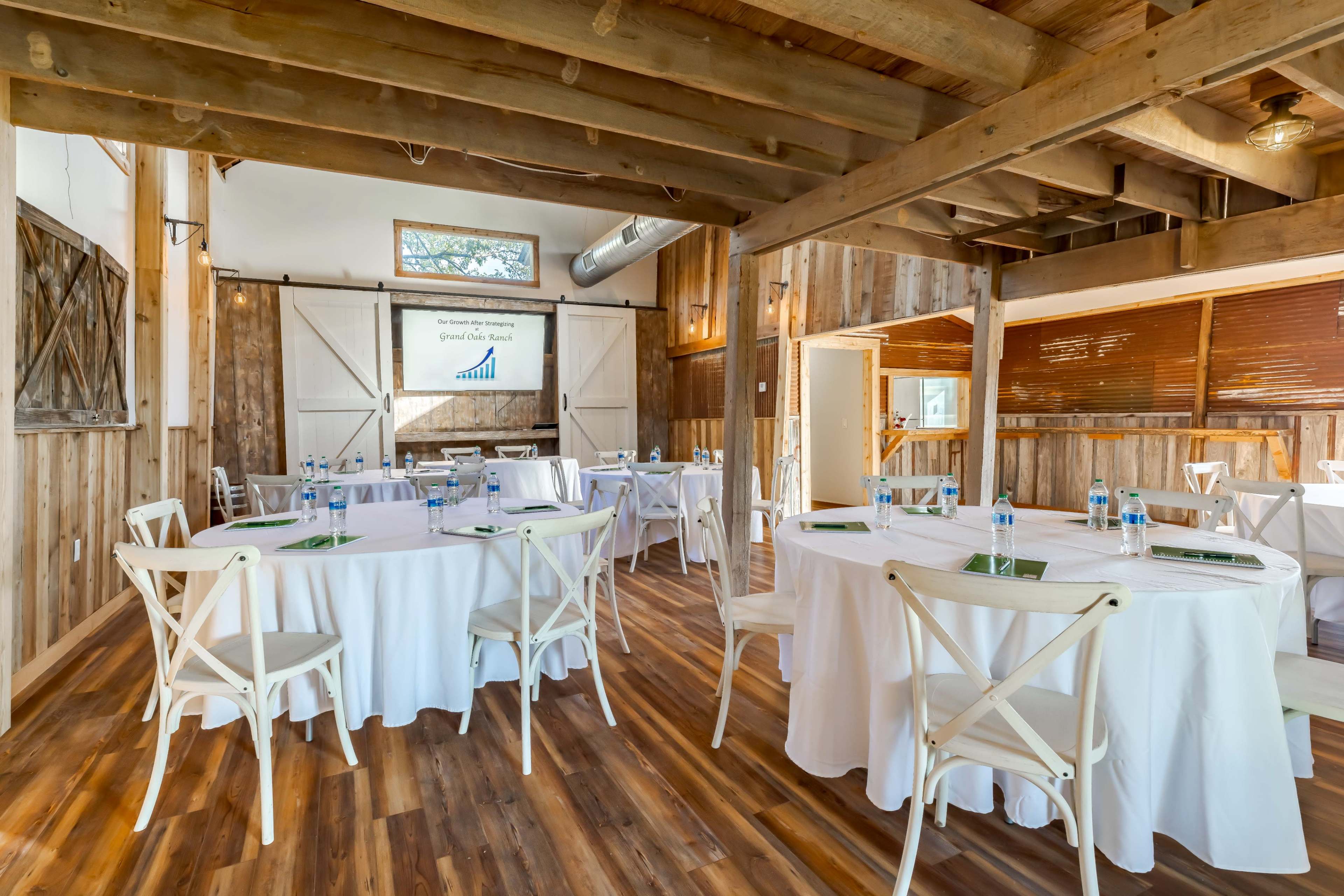 A rustic meeting room with round tables covered in white tablecloths, arranged for a presentation.
