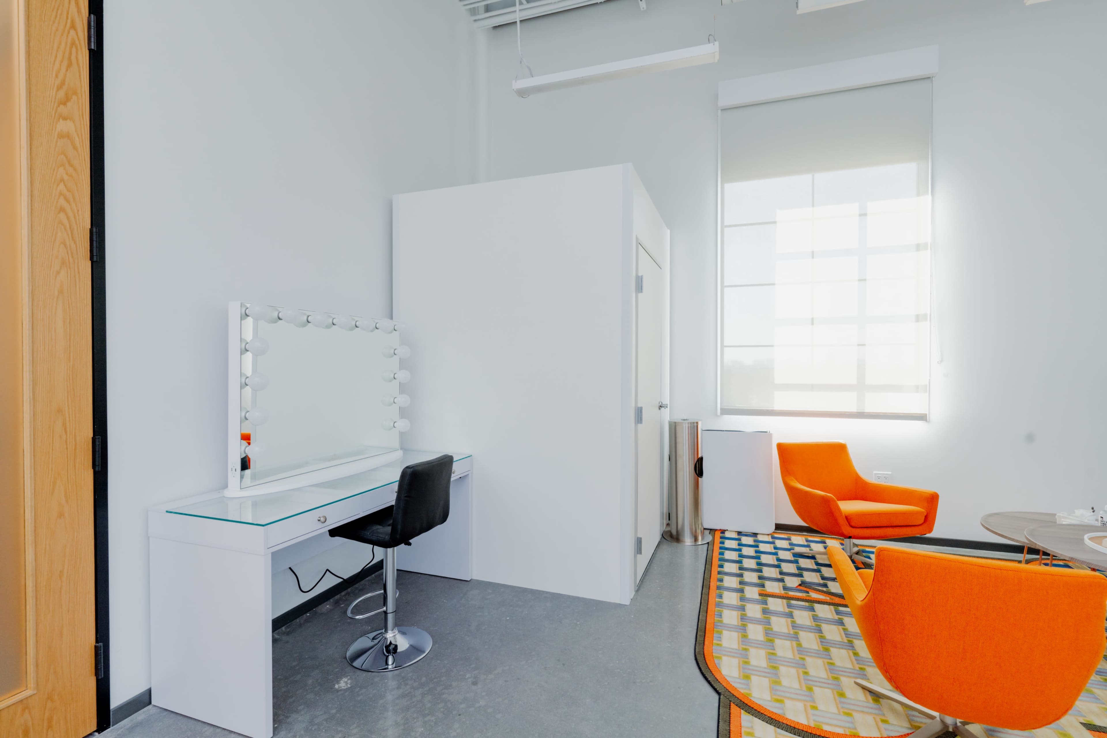 A modern vanity area with a large mirror, a black stool, bright orange armchairs, and a window letting in natural light.
