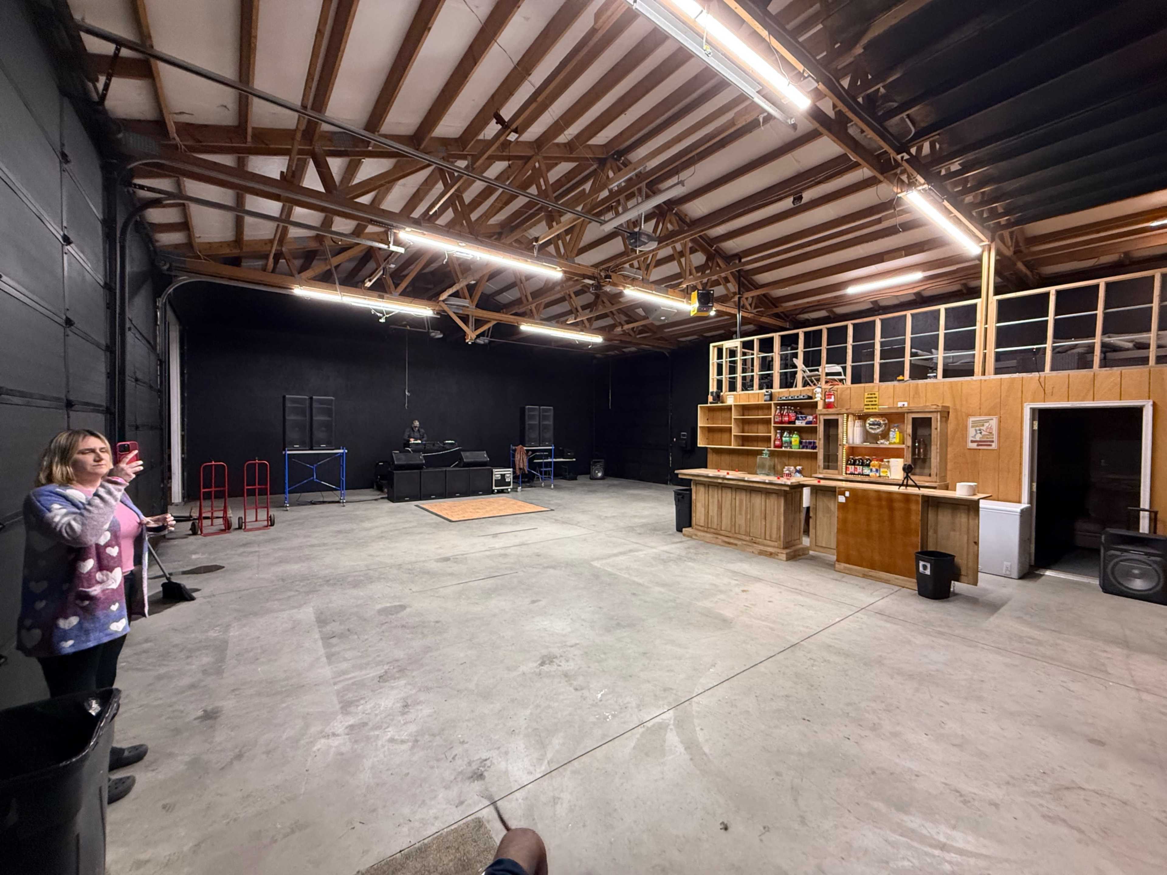 The image shows a large, empty warehouse space with exposed wooden beams, a bar area on one side, and a DJ setup in the background.