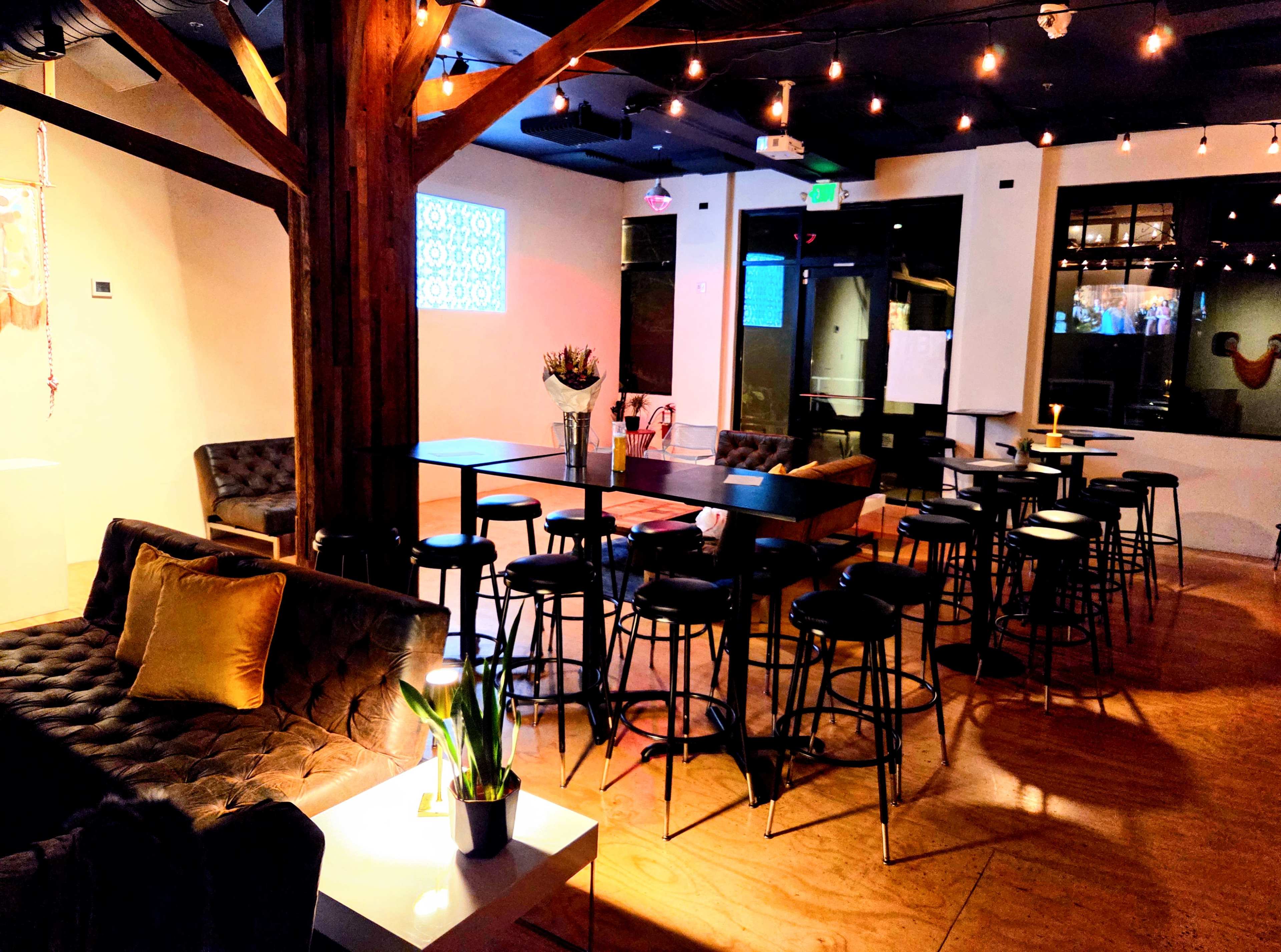 The image shows a cozy indoor space featuring a mix of high-top tables and lounge seating, illuminated by string lights and accented with plants.