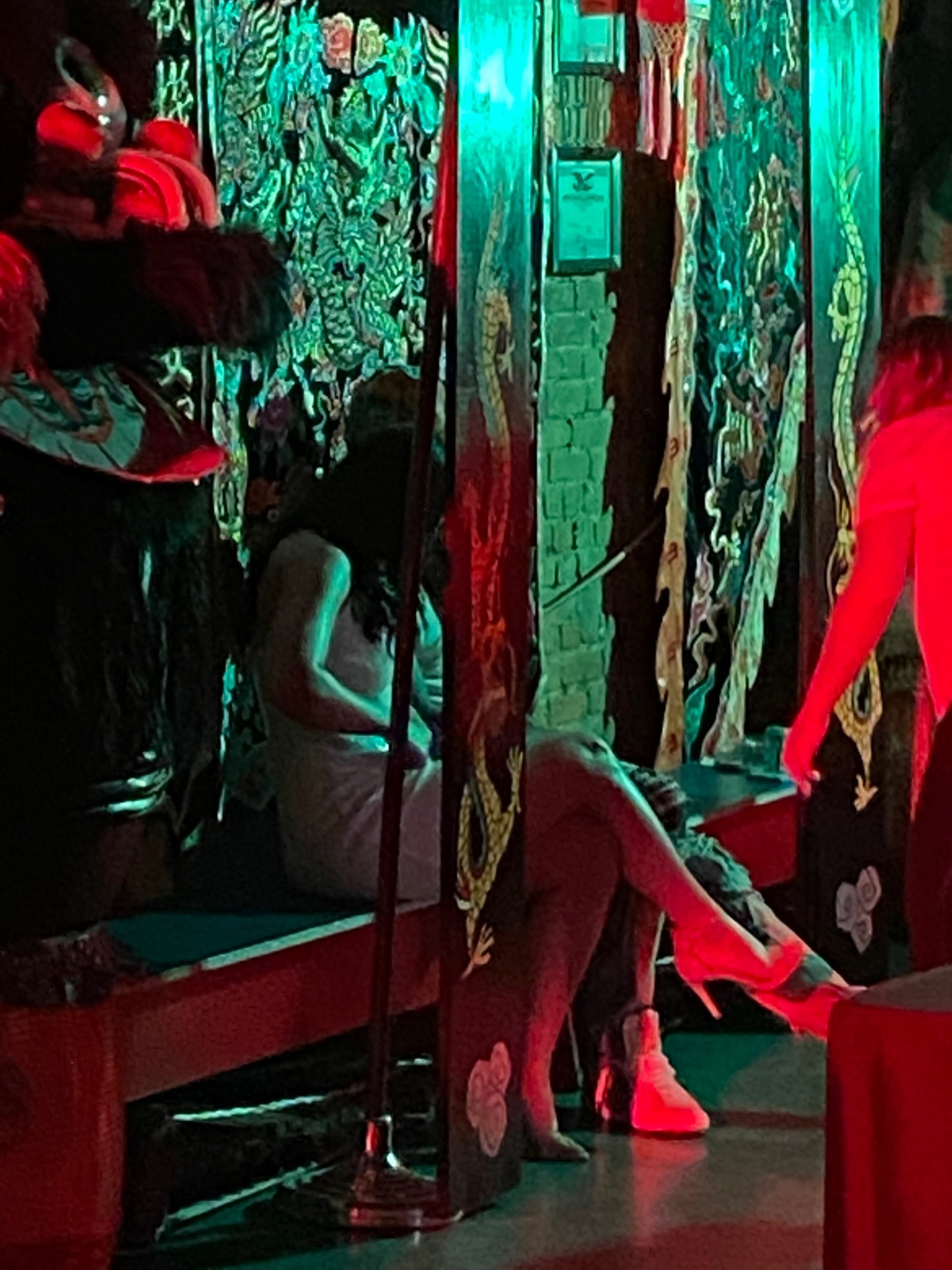 The image shows a dimly lit interior with patterned fabric hanging and two people sitting on a bench, one in a white outfit and the other in red.