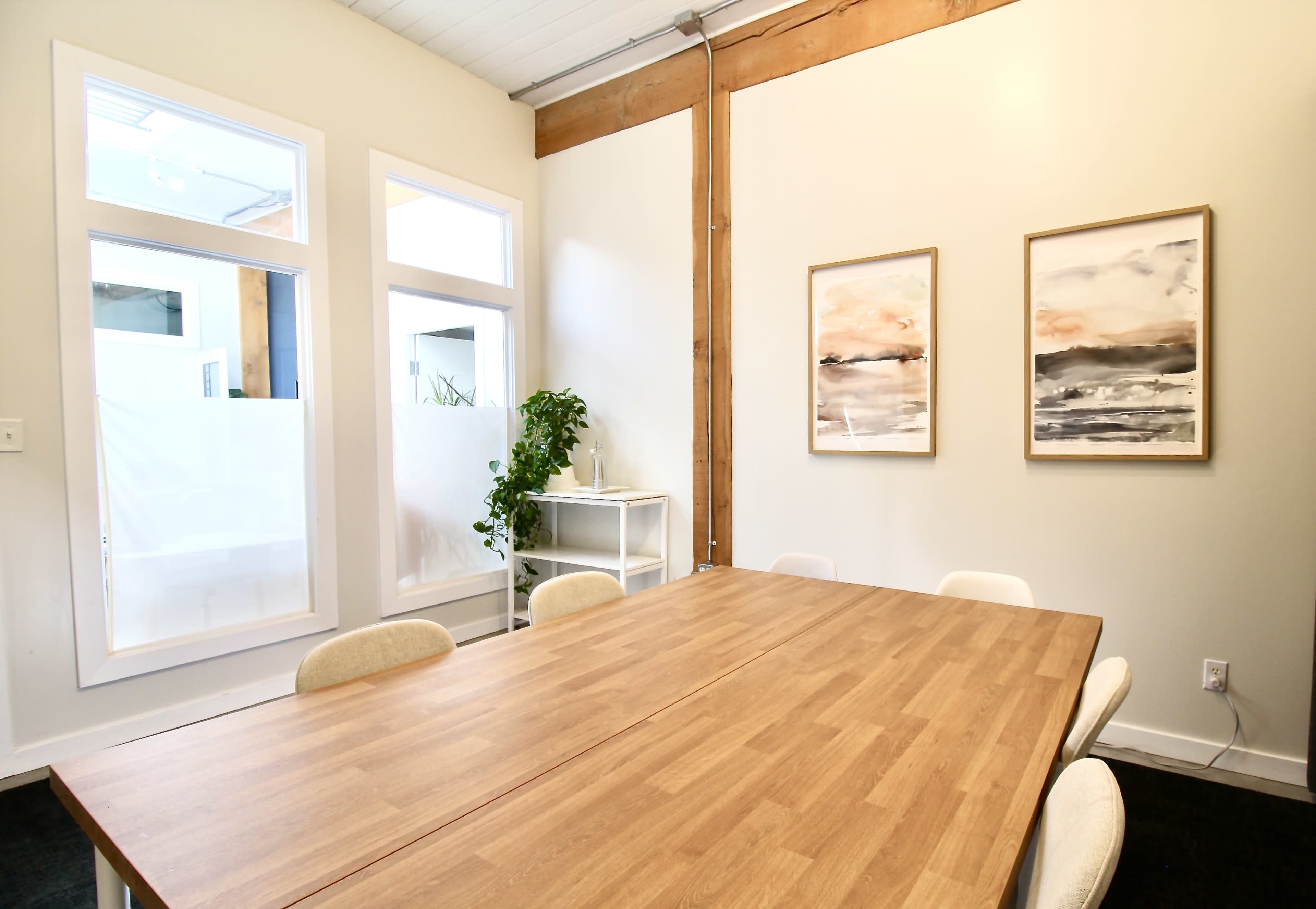 A wooden conference table is set in a bright meeting room adorned with two framed artworks on the wall and a potted plant nearby.