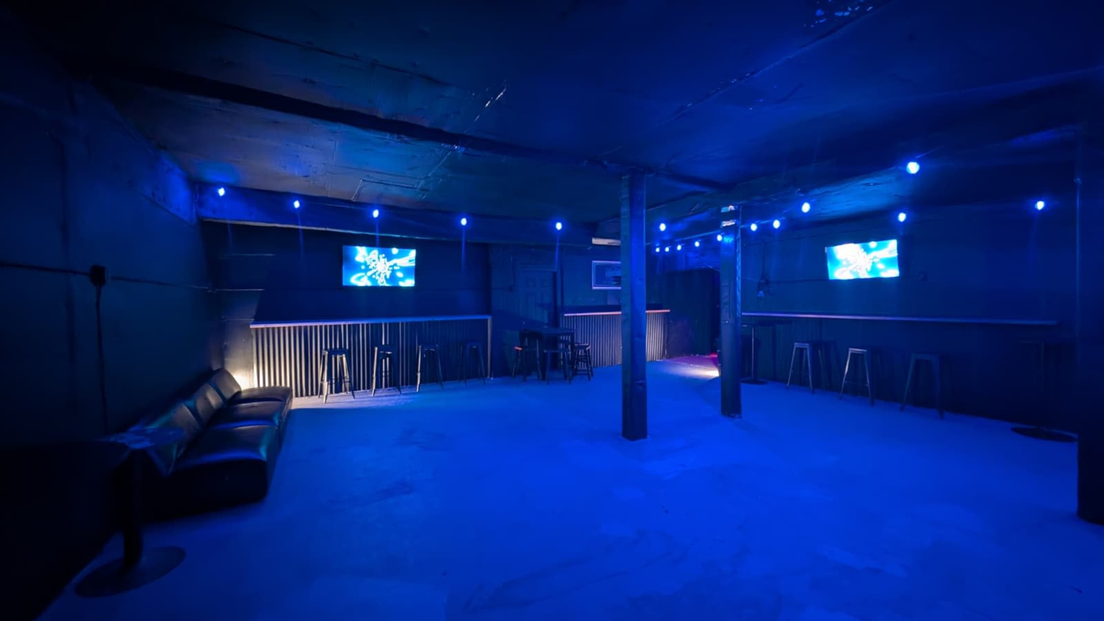 The image shows an empty, dimly lit room with blue lighting, featuring a couch, tables, and screens mounted on the walls.