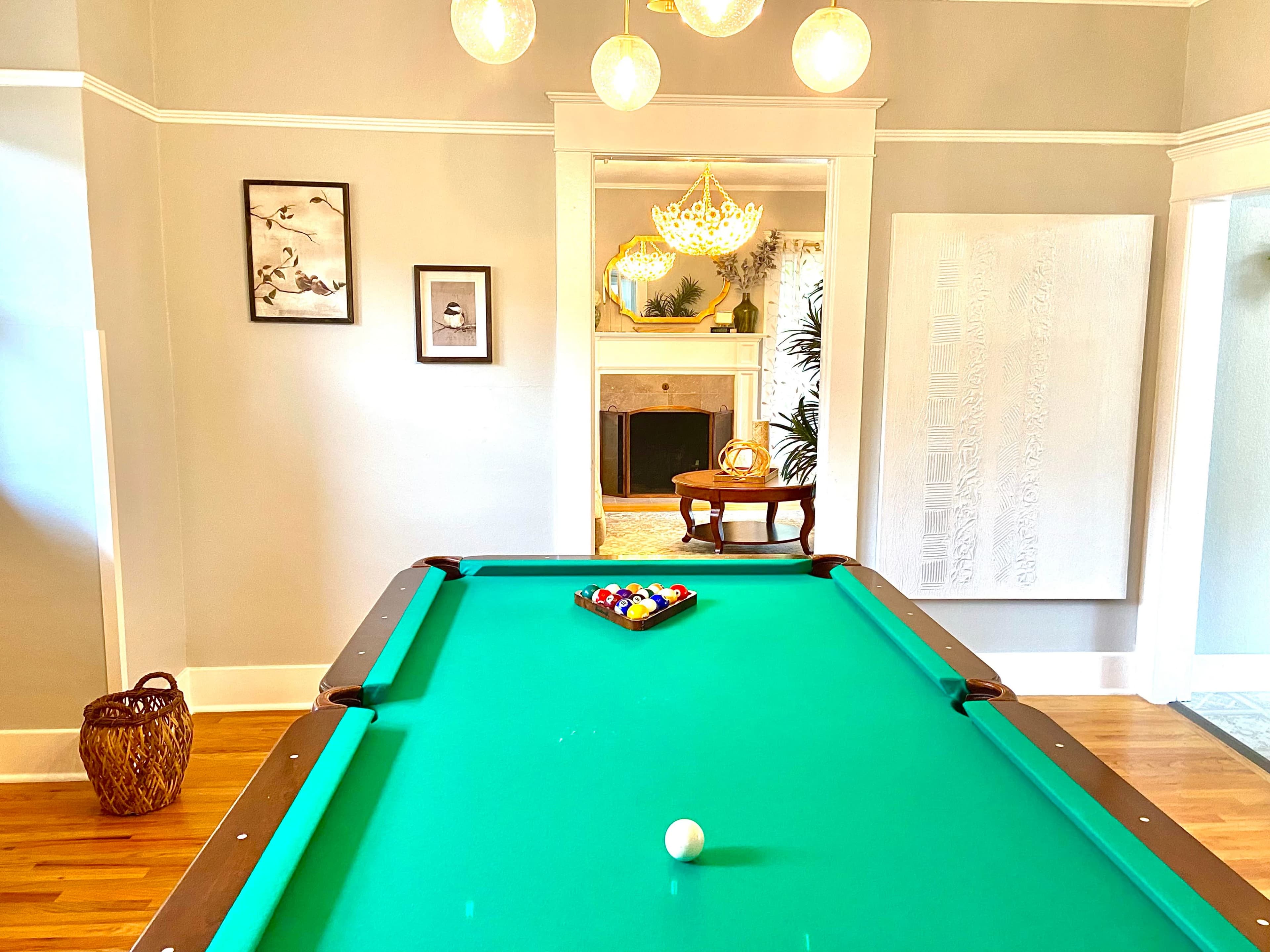 A green pool table with a triangle of colored balls sits in a well-lit room featuring artwork on the walls and a chandelier overhead.