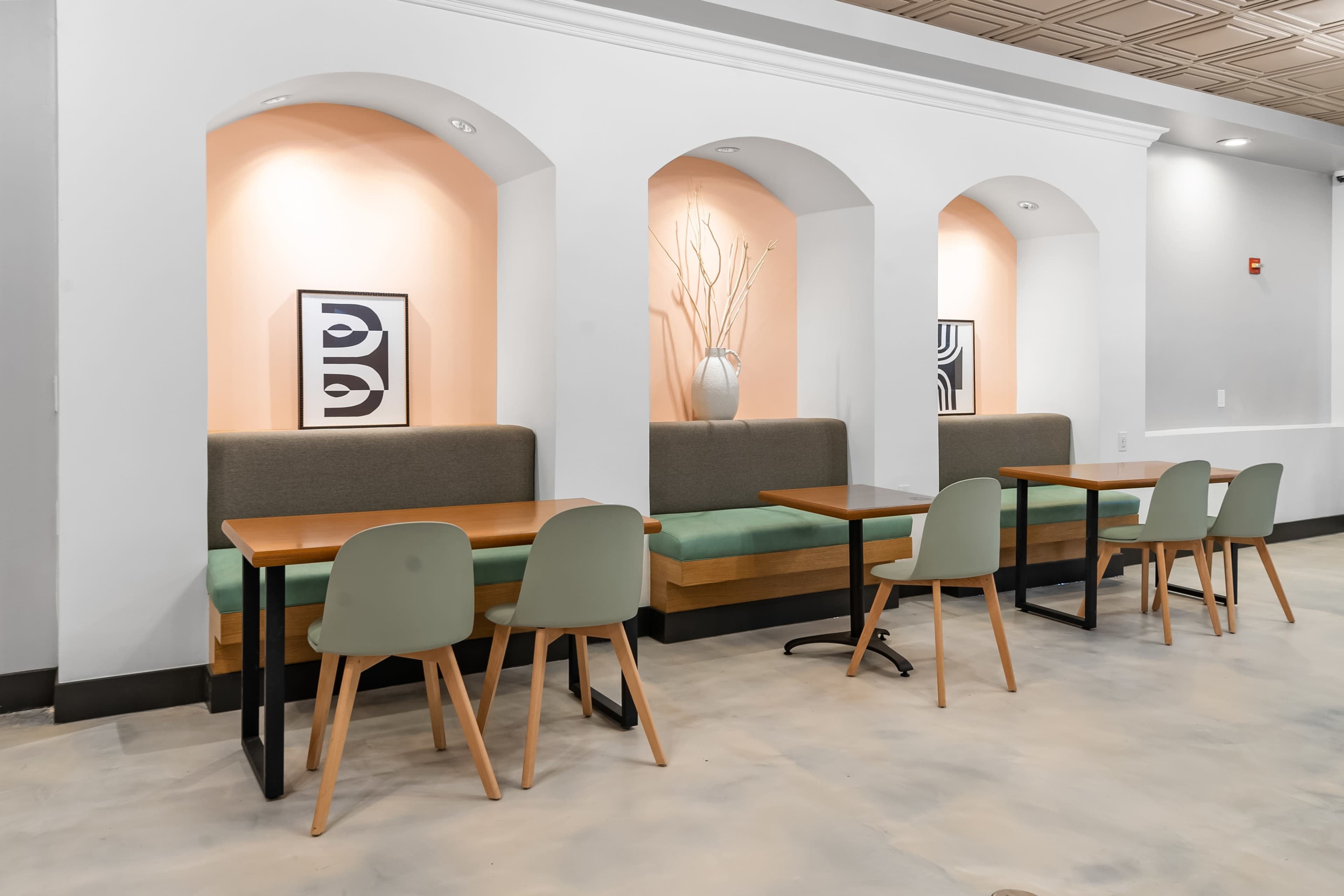 A light-colored cafe interior with several seating areas featuring green cushioned benches, wooden tables, and modern chairs arranged against a white wall with arched niches.