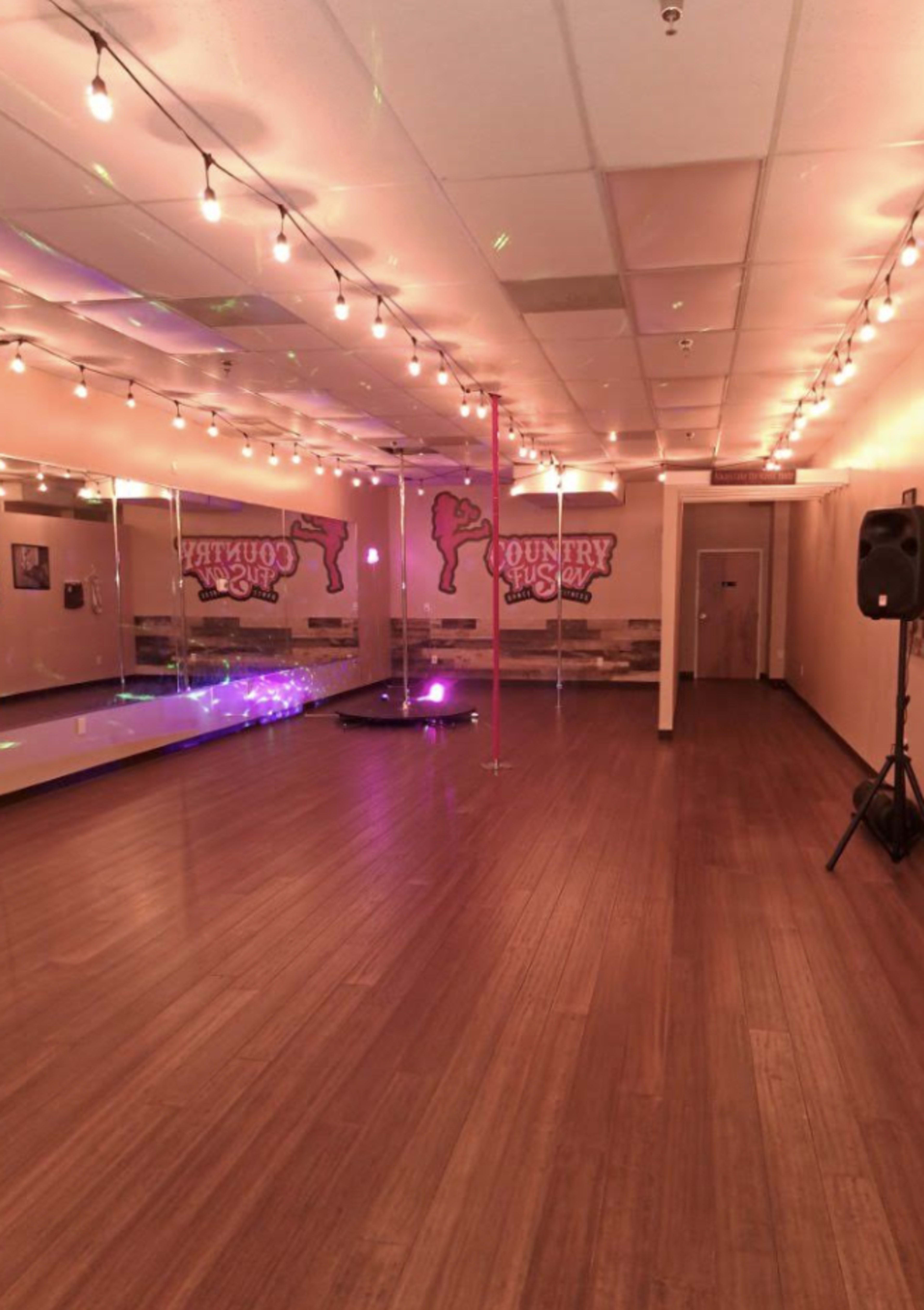 A dance studio with a polished wooden floor, mirrors on one wall, a pole in the center, and string lights overhead.