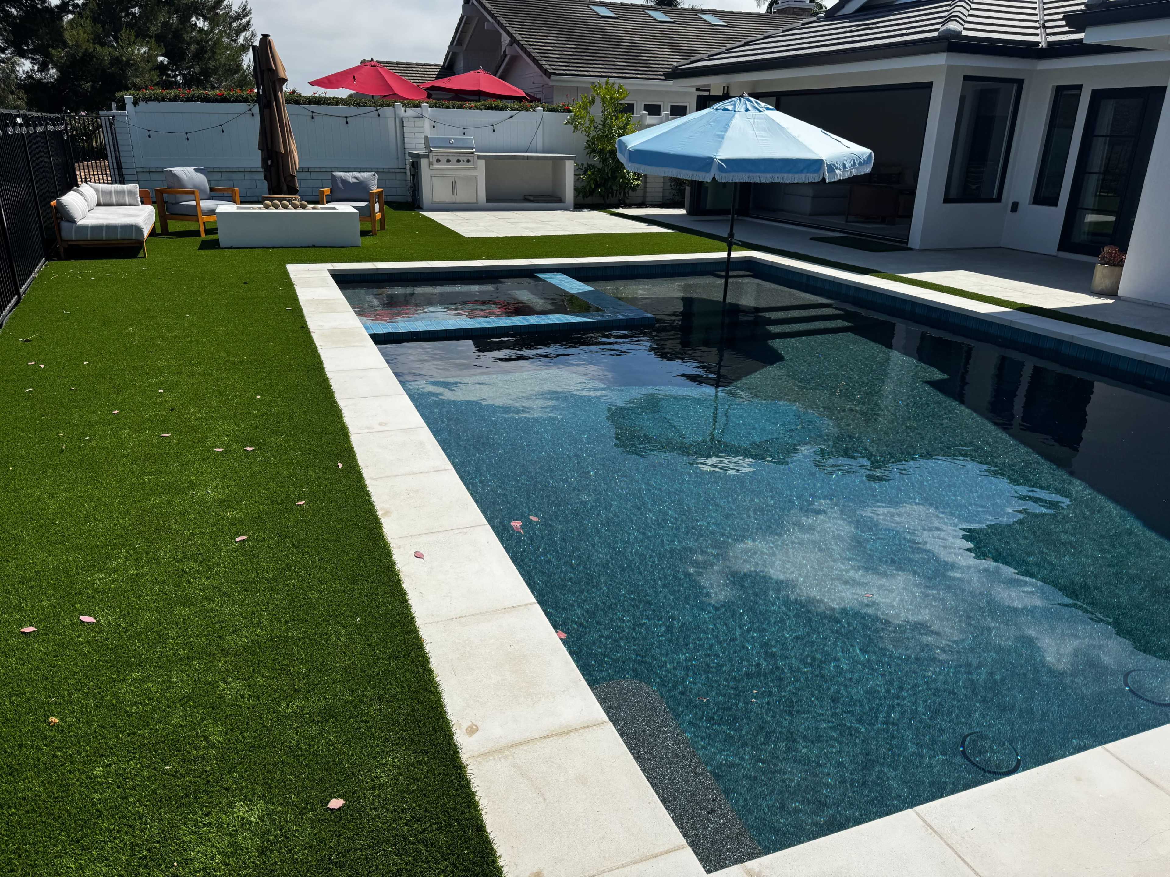 The image shows a modern backyard with a swimming pool surrounded by artificial grass, lounge chairs, an umbrella, and a barbecue area.
