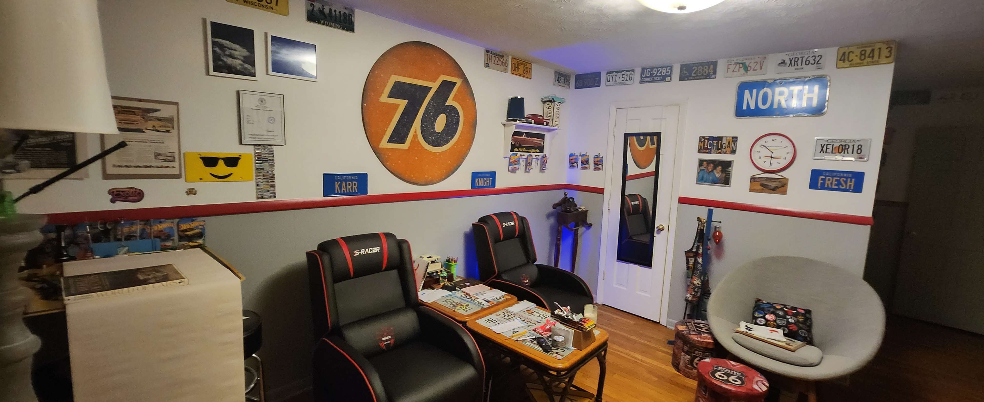 The image depicts a cozy room featuring two gaming chairs, decorative wall art including a large 76 logo, and various vintage license plates and photographs adorning the walls.