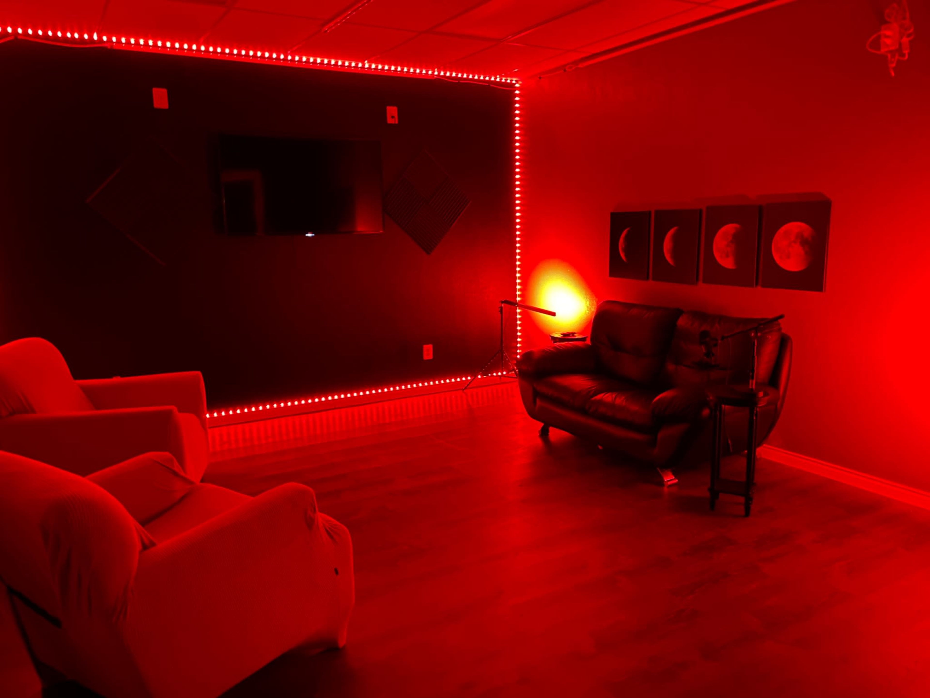 The image depicts a room with red walls, a black sofa, two armchairs, a television mounted on the wall, and decorative artwork featuring moons.