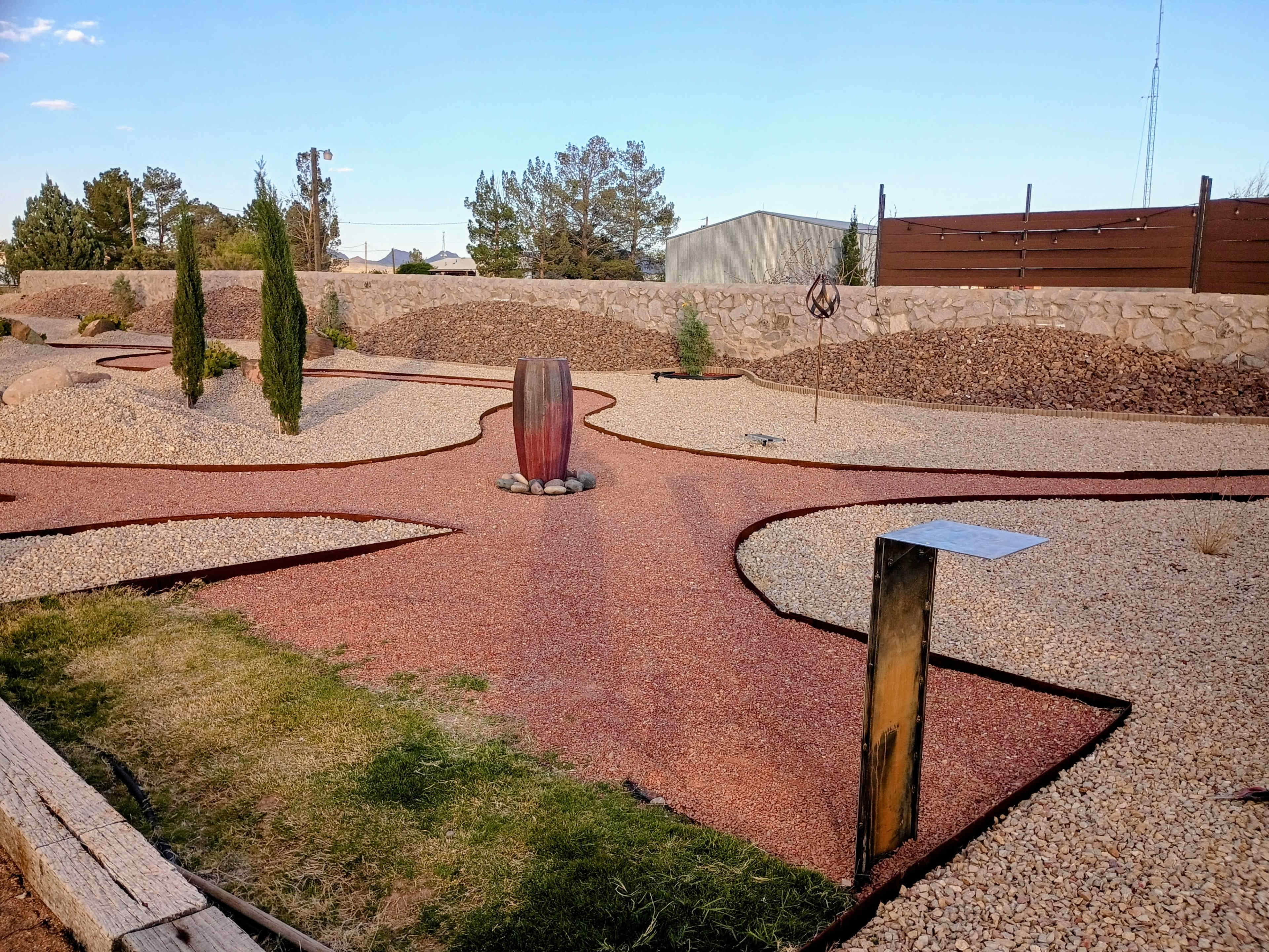 The image depicts a landscaped garden featuring a winding path lined with red gravel, surrounded by decorative rocks and small trees.