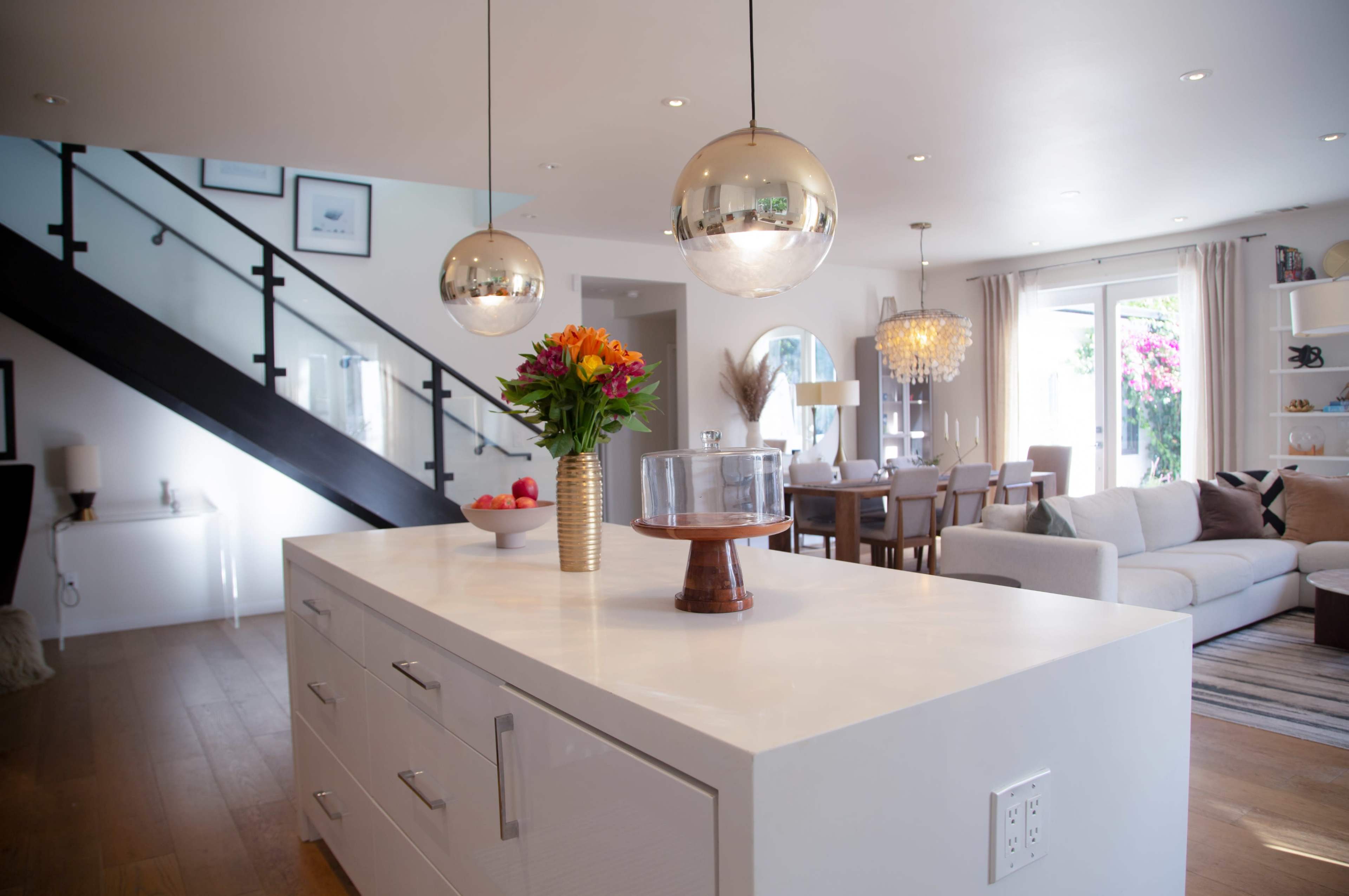 A modern kitchen features a central island with a vase of flowers, pendant lights above, and a view of a dining area in the background.