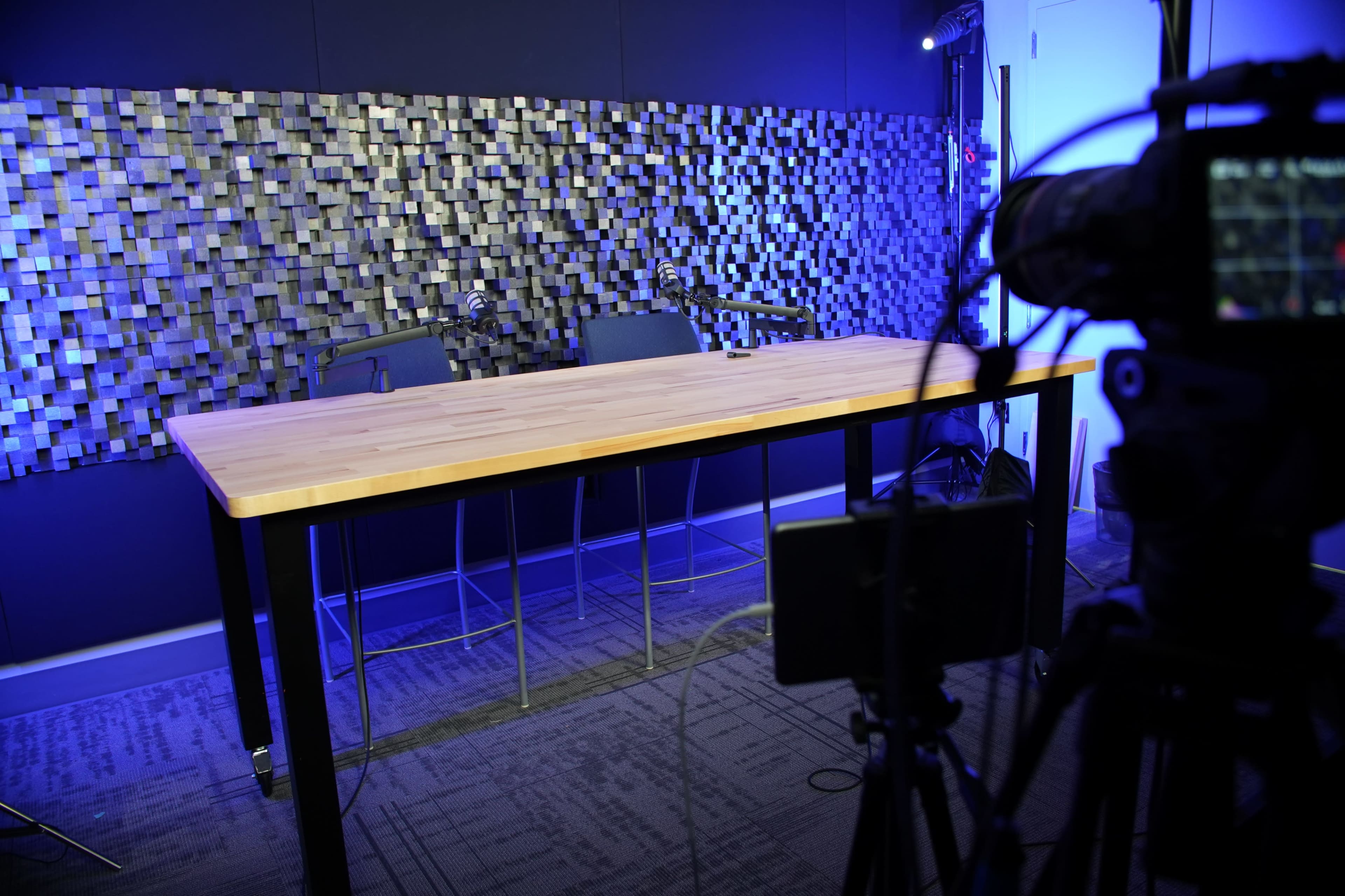 The image shows a modern recording studio setup with a large wooden table, two empty chairs, and a textured wall behind, illuminated by blue lighting.
