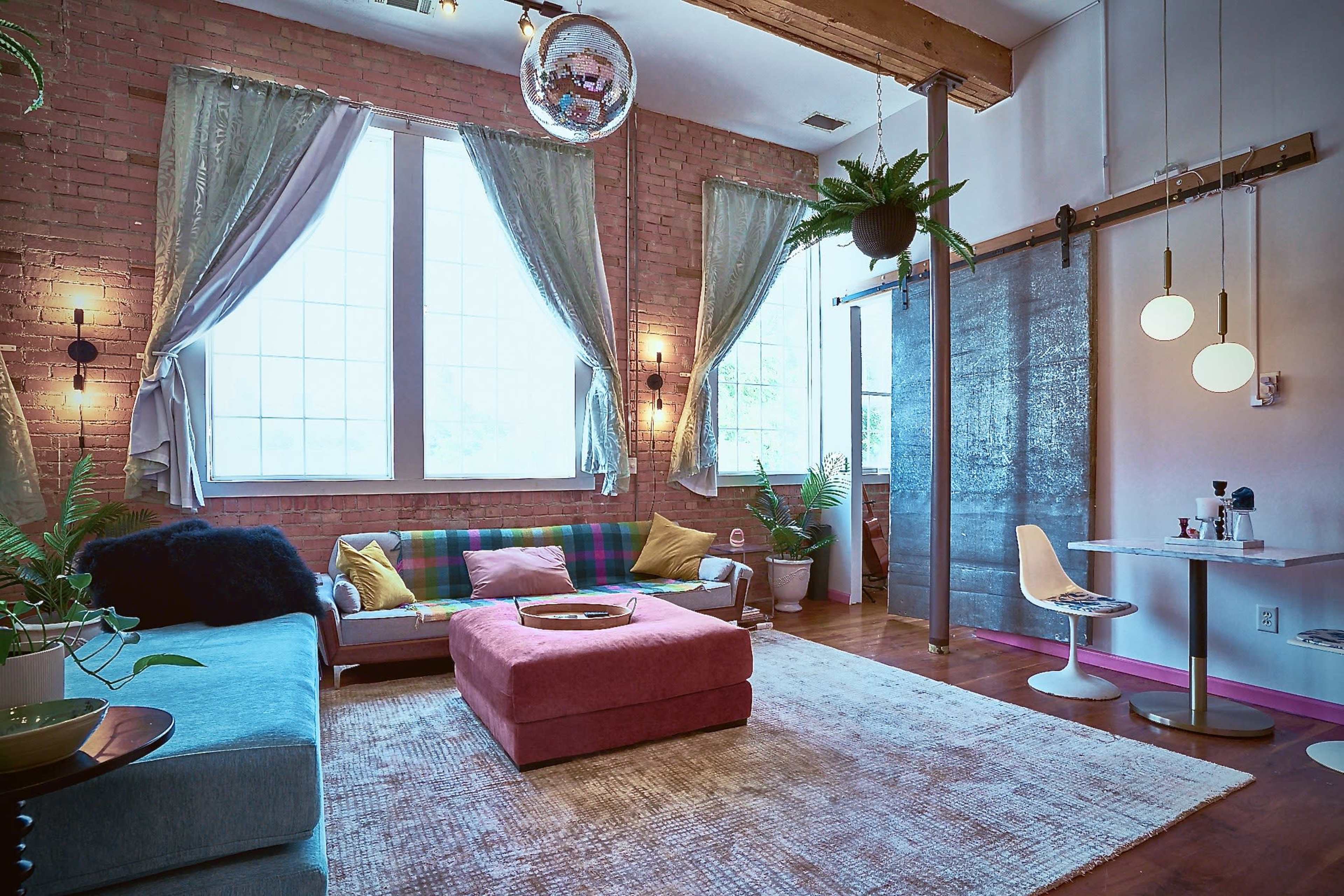 A colorful living room with exposed brick walls, large windows, and a mix of modern and vintage furniture.