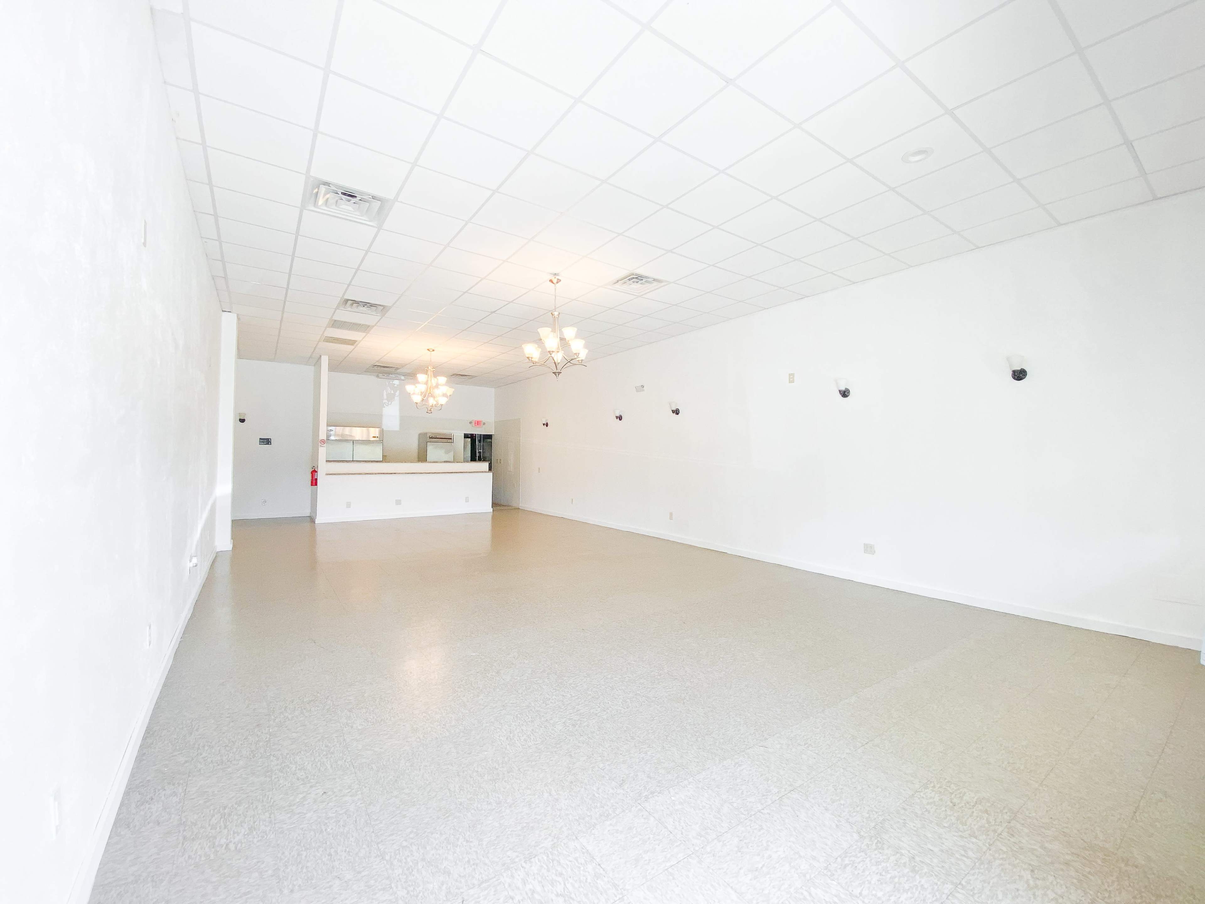 The image shows a spacious, empty room with white walls and a tile floor, featuring a counter area and several light fixtures.