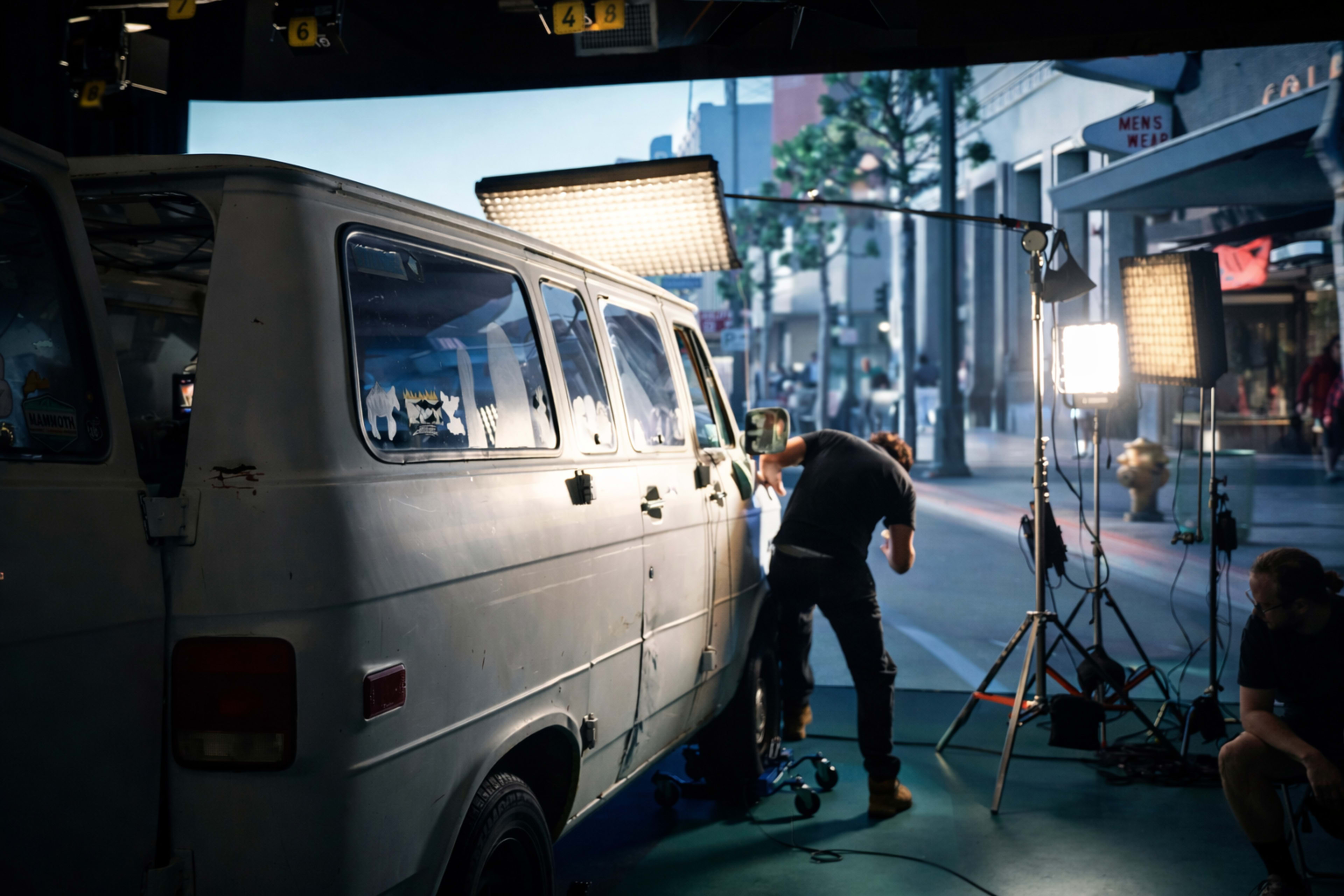 A crew member is preparing a white van in front of a large backdrop depicting a street scene, with studio lights set up for filming.