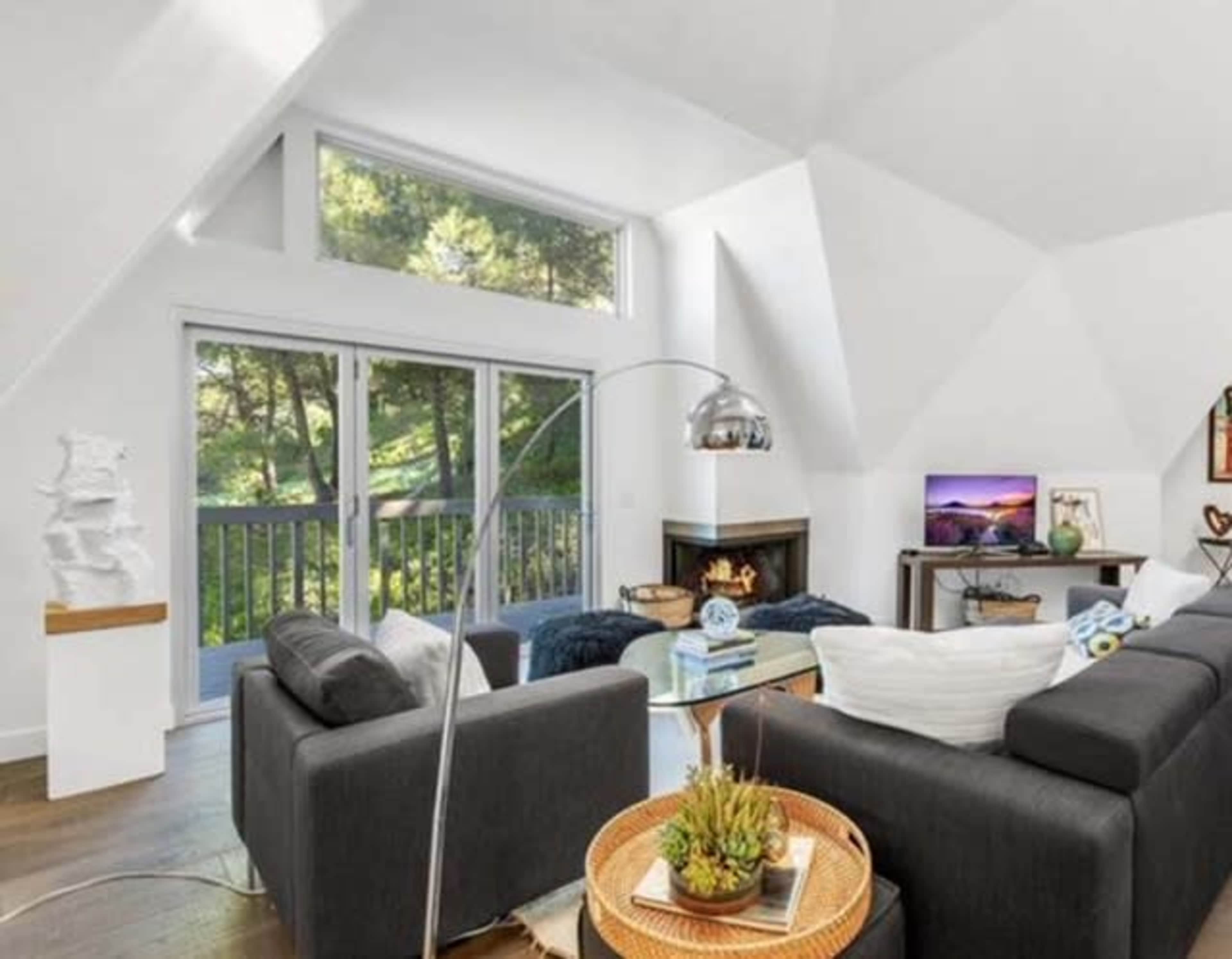 A modern living room with a triangular ceiling features a gray sectional sofa, a glass coffee table, and large windows overlooking greenery.