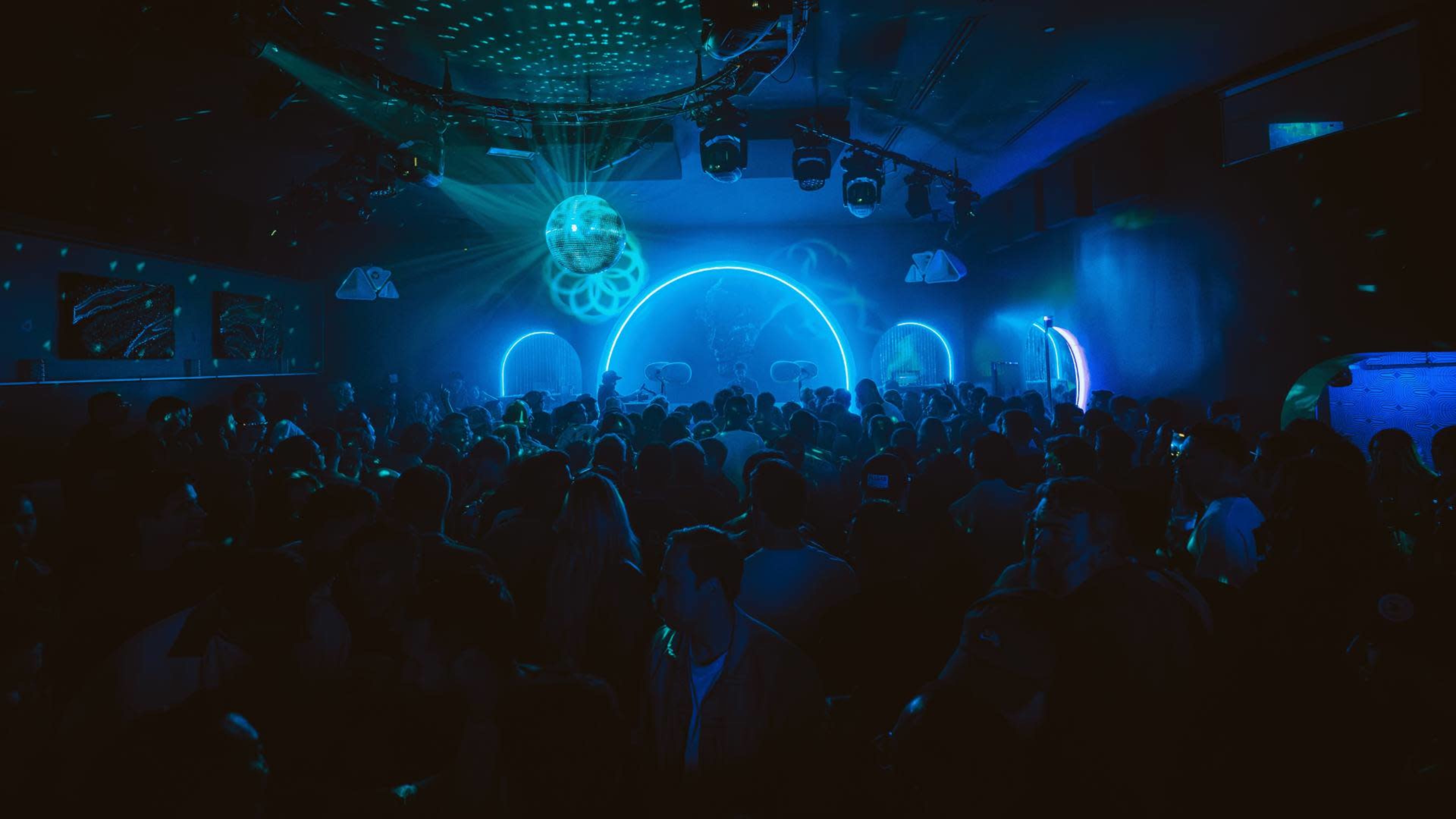 A crowded nightclub features vibrant lighting and a large dance area filled with people.