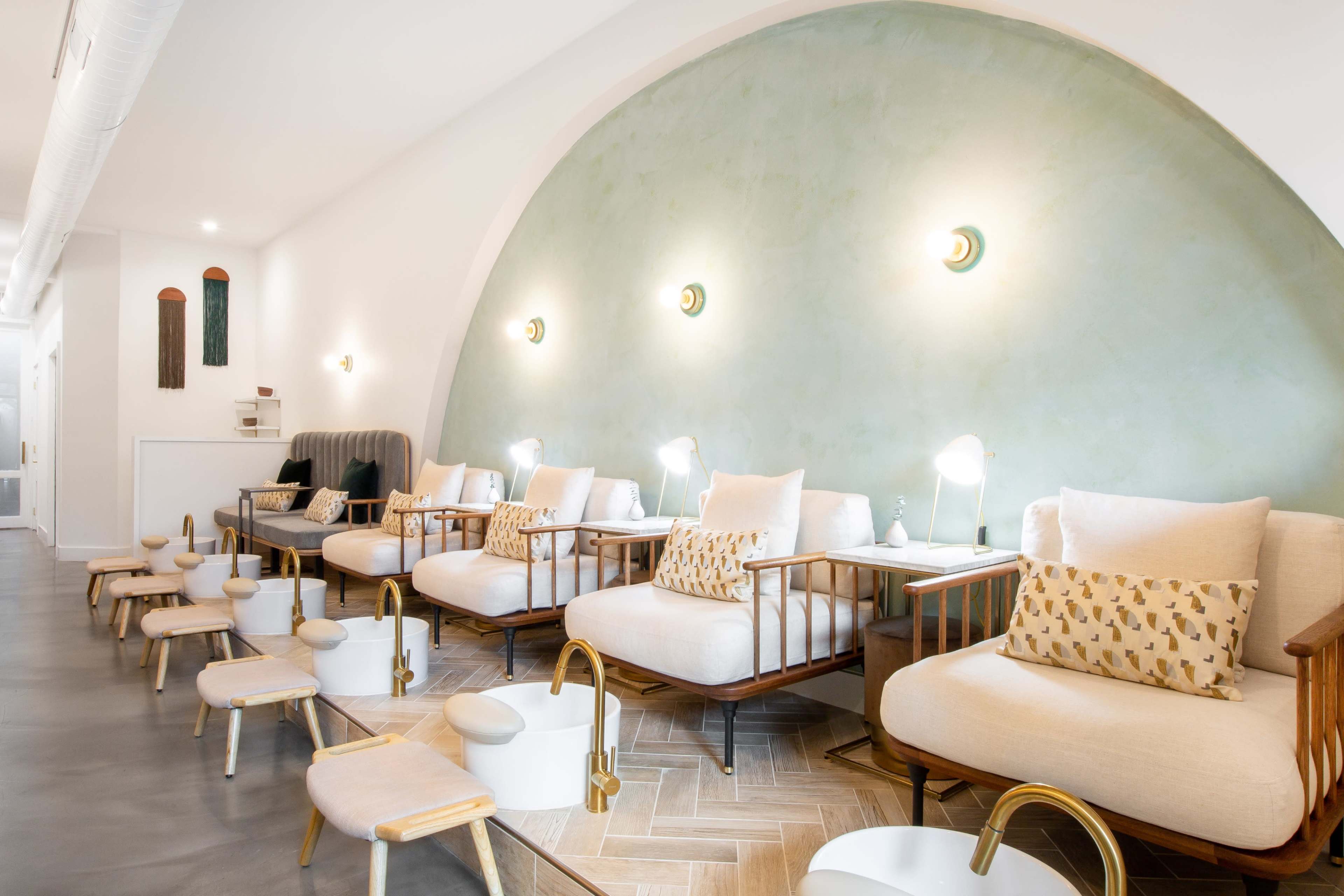 A modern spa interior featuring a row of cushioned chairs next to small foot baths, all under a curved green wall with soft lighting.