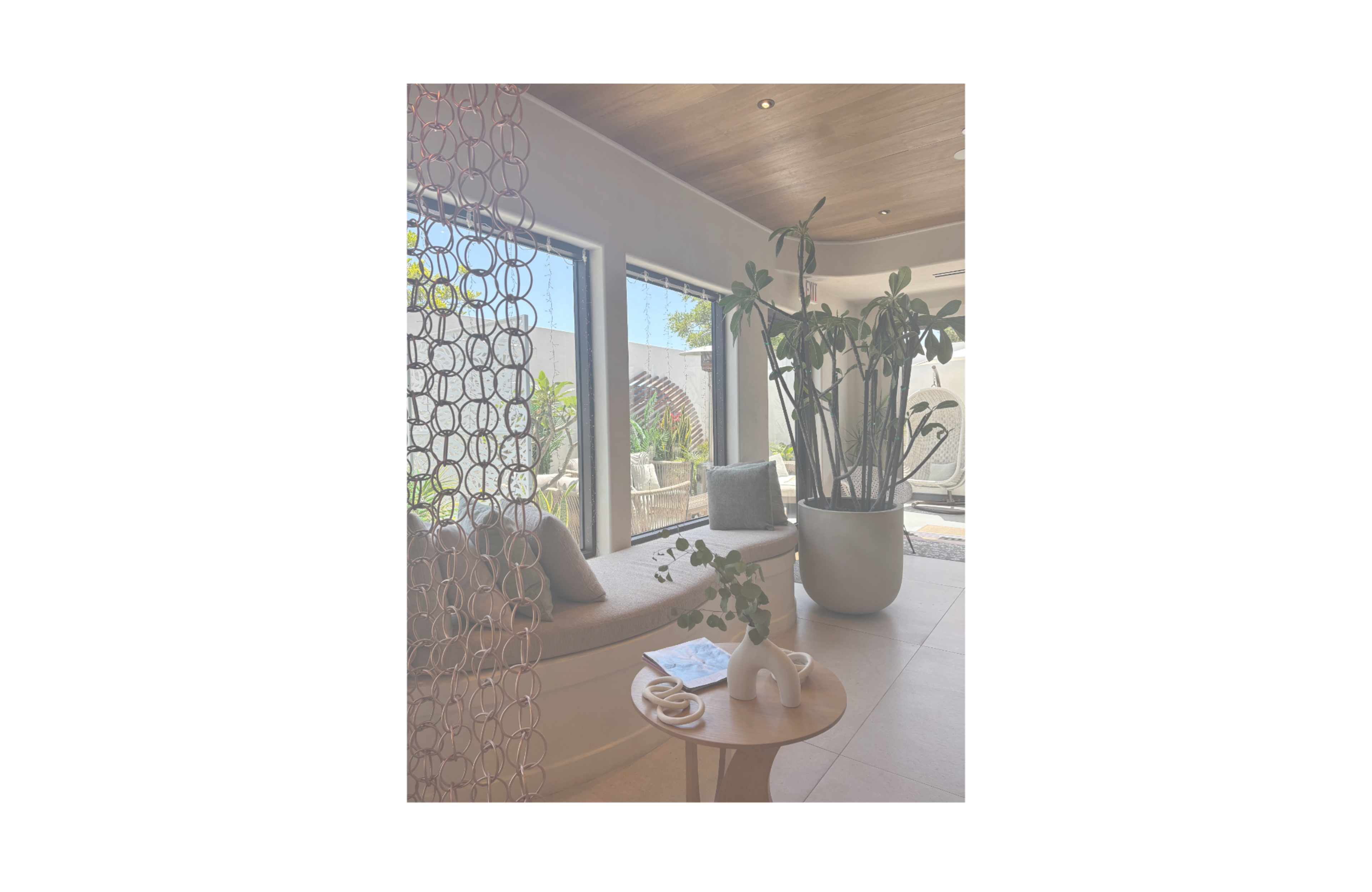 The image shows a modern indoor space with a cushioned seating area and large windows featuring plants and a wooden ceiling.