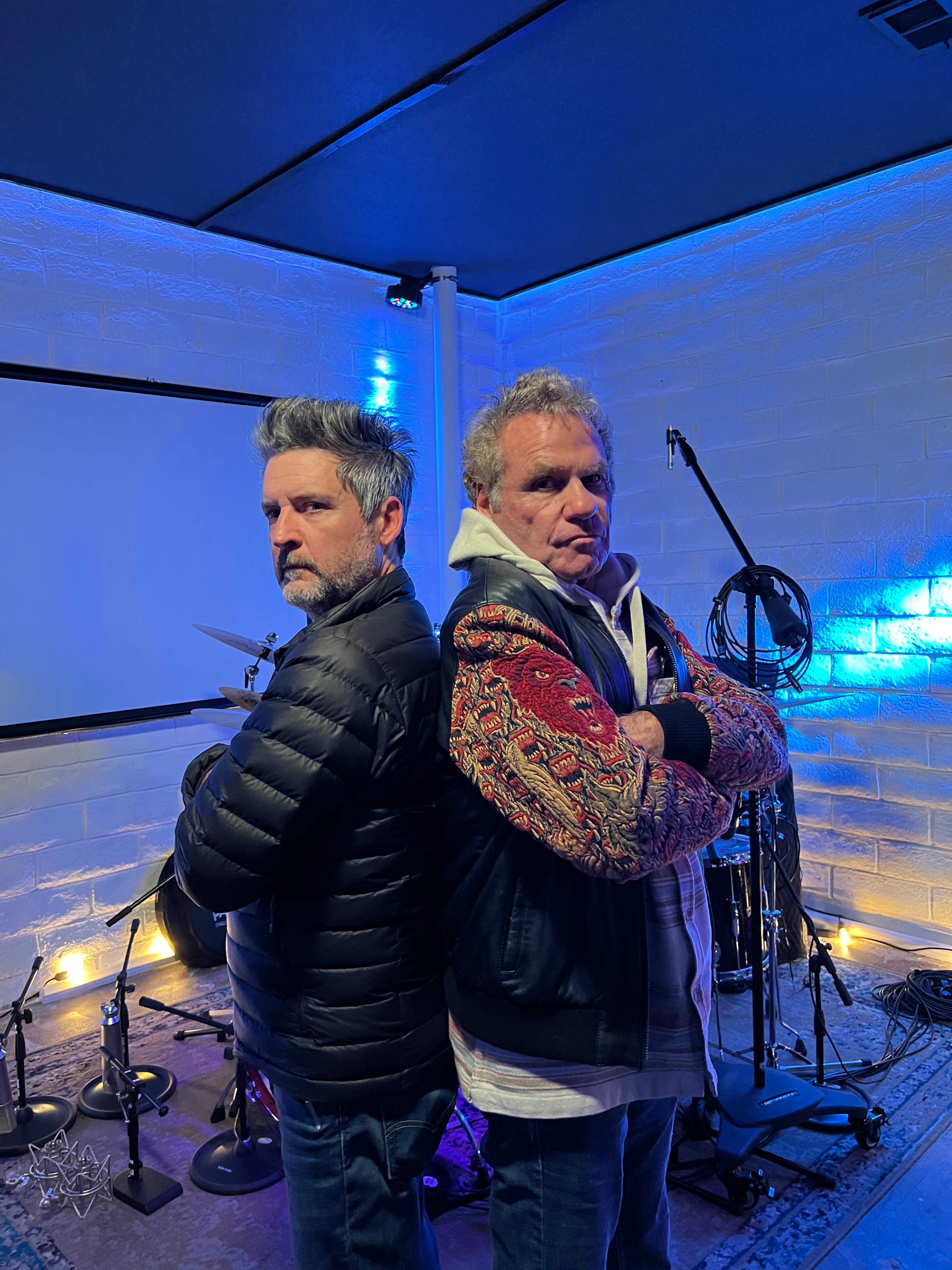 Two men stand back to back in a recording studio, each with arms crossed, surrounded by musical instruments and blue lighting.