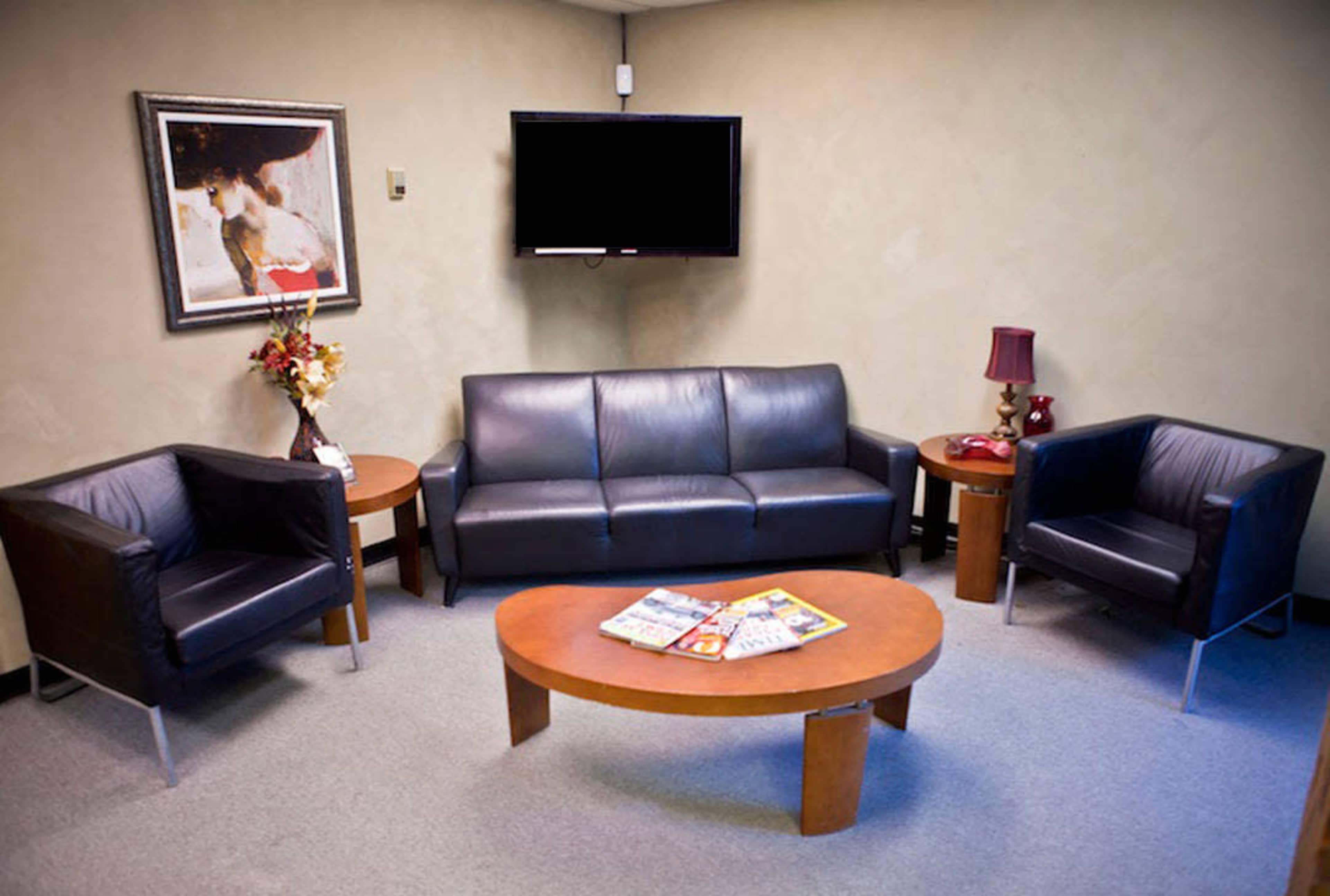 A waiting room features a black leather sofa and two matching chairs positioned around a round coffee table, with a television mounted on the wall and a decorative painting nearby.