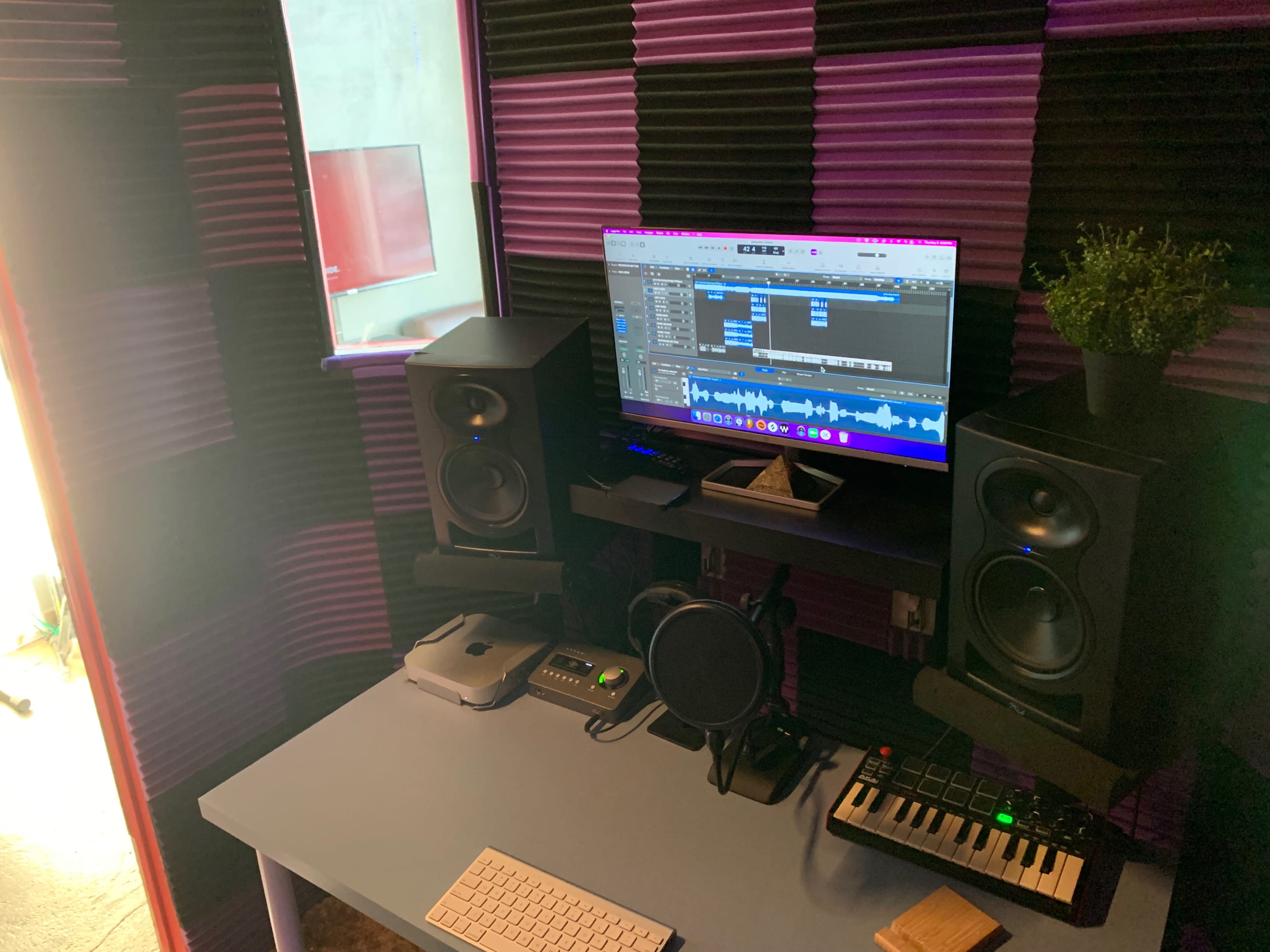 A music production setup featuring a computer monitor displaying audio software, two studio speakers on either side, a microphone, a small keyboard, and soundproofing panels on the walls.