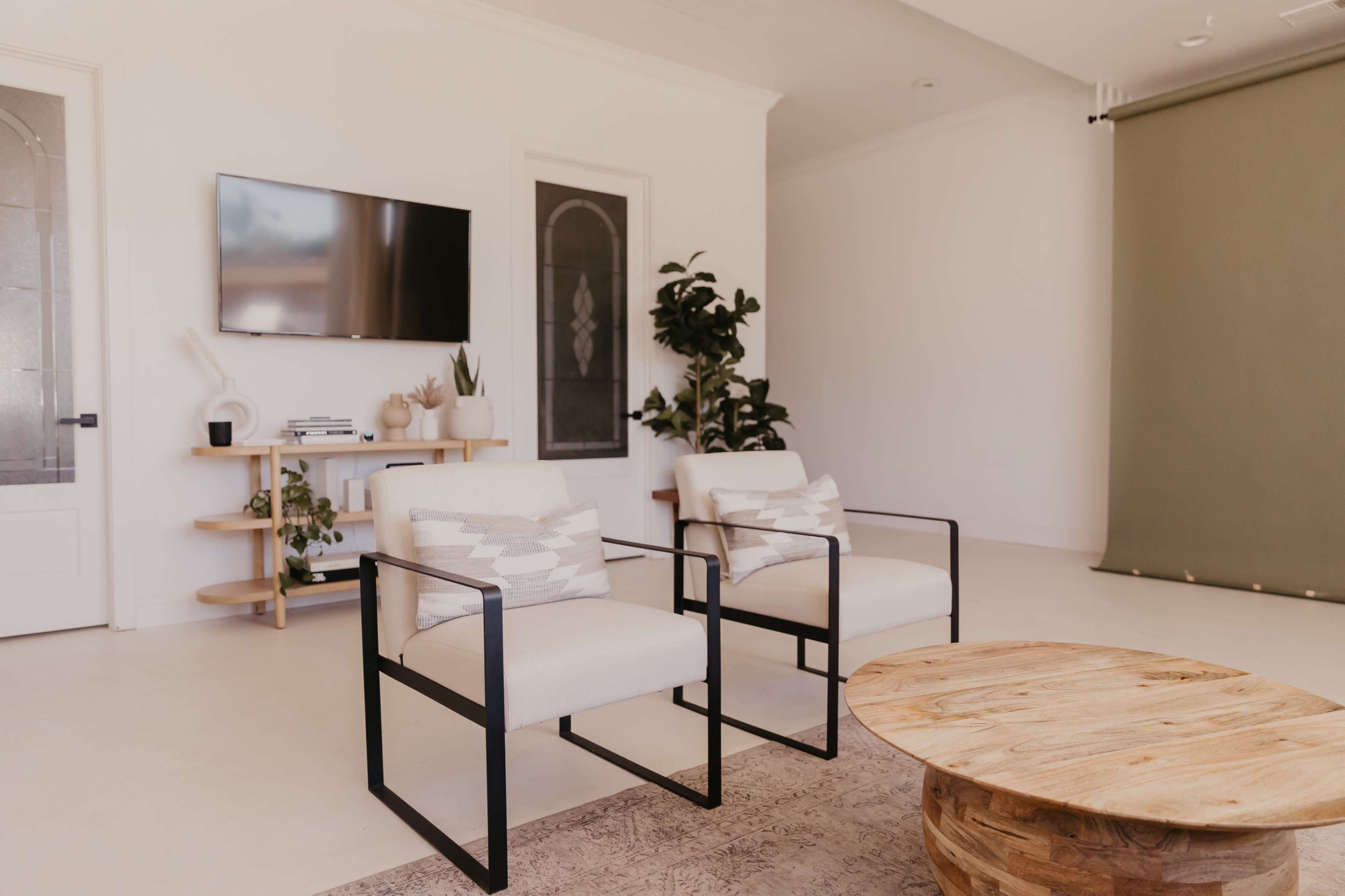 A modern living room with two white armchairs, a round wooden coffee table, a television mounted on the wall, and a corner plant beside a green backdrop.