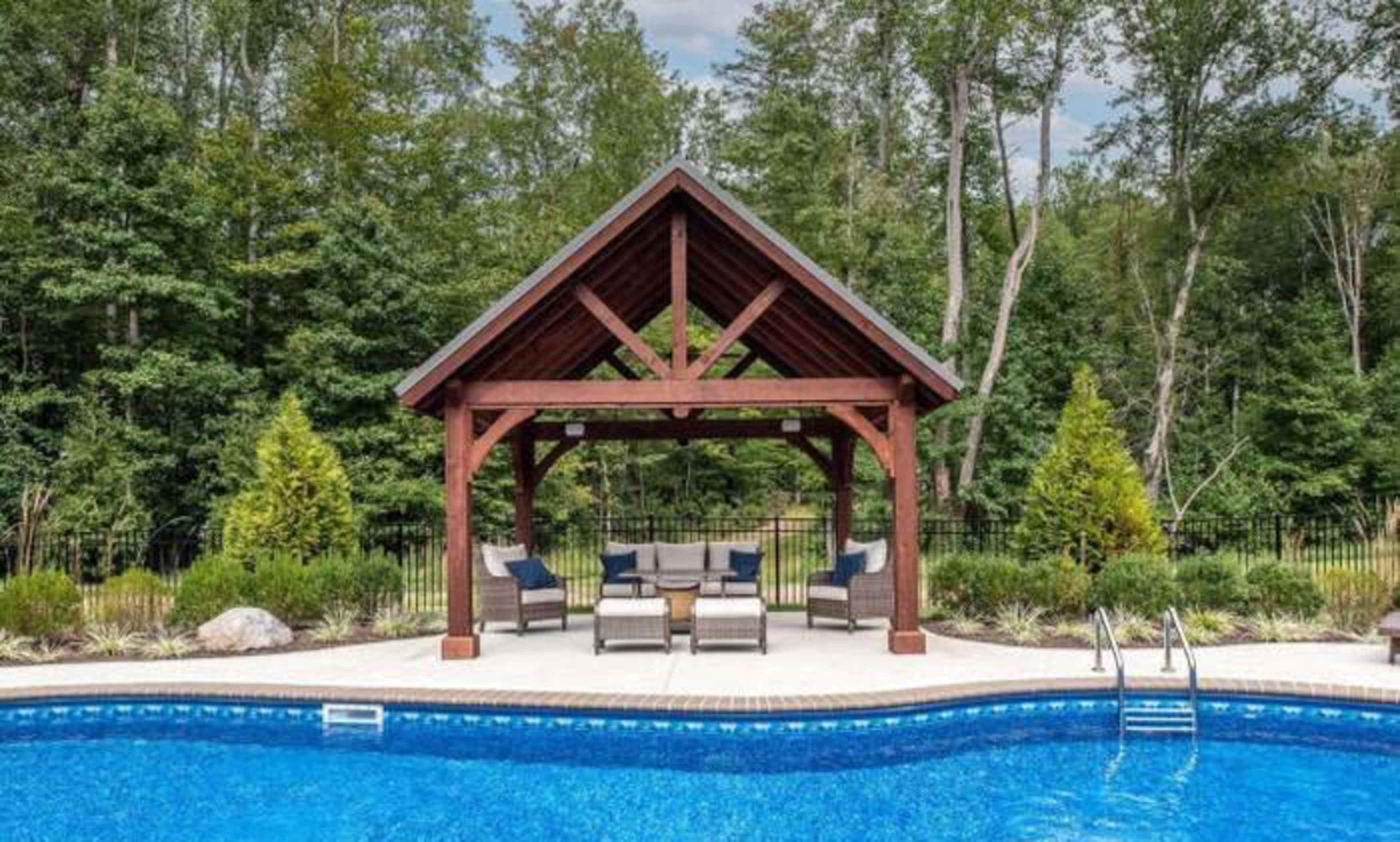 A wooden gazebo with seating overlooks a blue swimming pool surrounded by greenery.