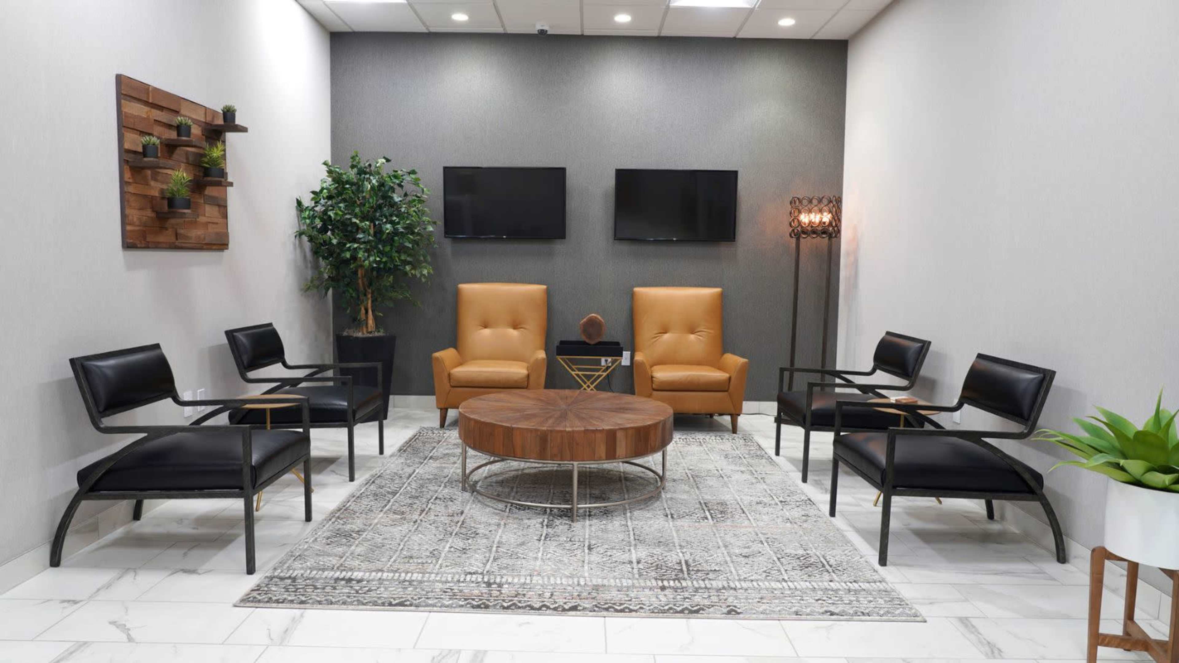 A modern waiting area features two orange armchairs, four black chairs, a round wooden coffee table, and a decorative rug, with potted plants and wall-mounted decor.