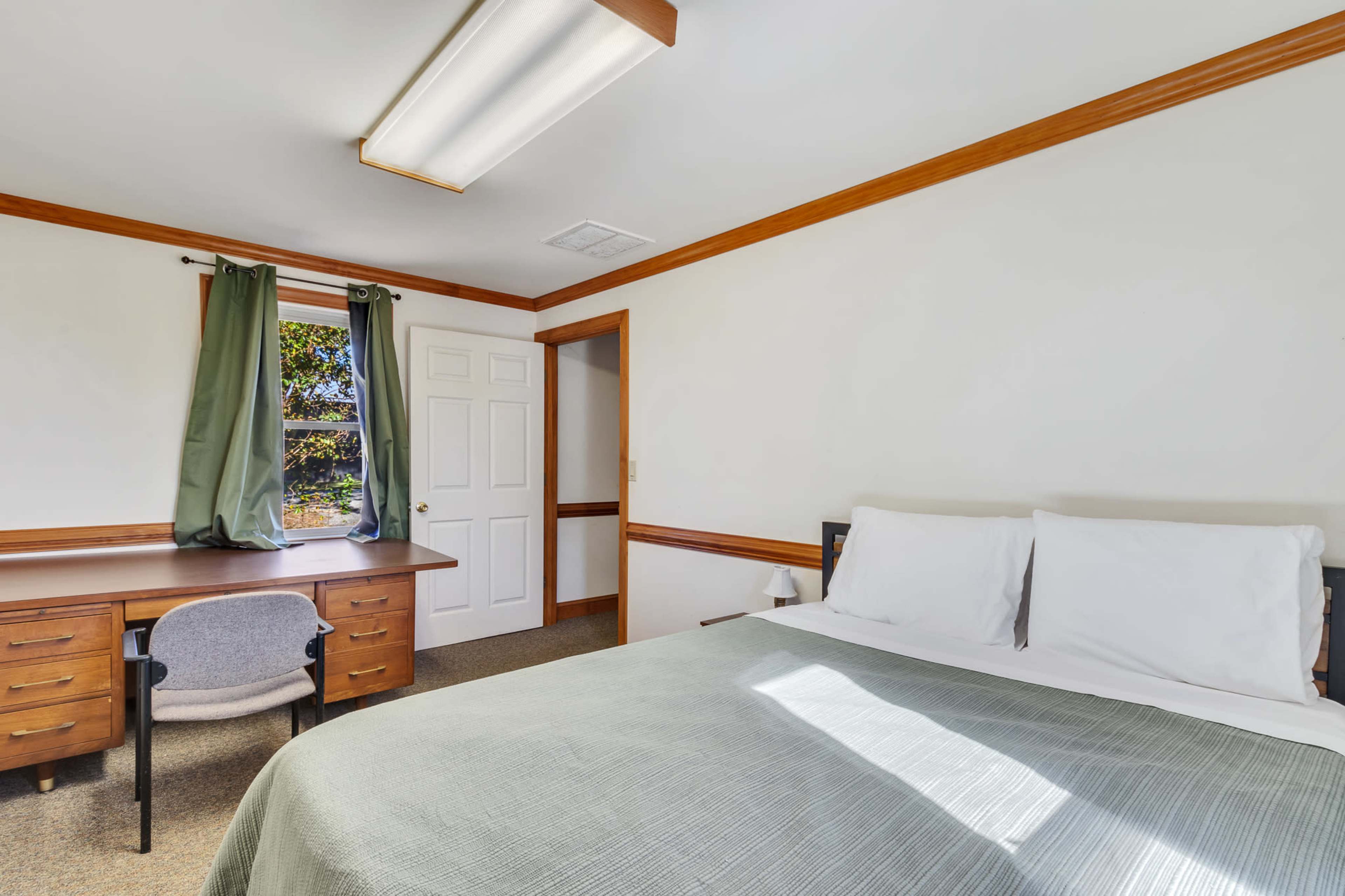 A simple bedroom features a bed with white linens, a wooden desk, a chair, and a window with green curtains.