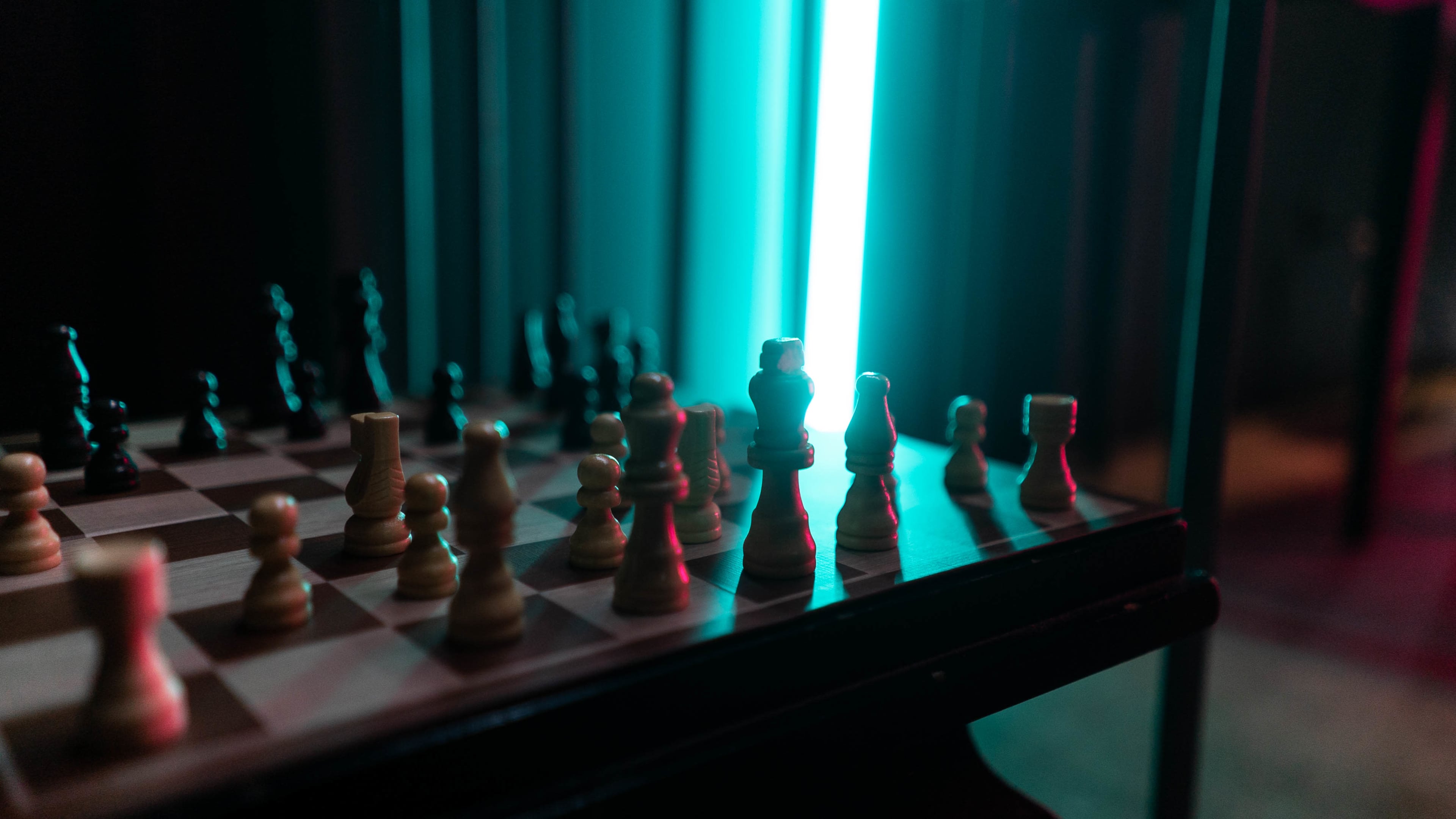 A chessboard is illuminated by a bright blue light, highlighting the wooden and black pieces in a dim setting.
