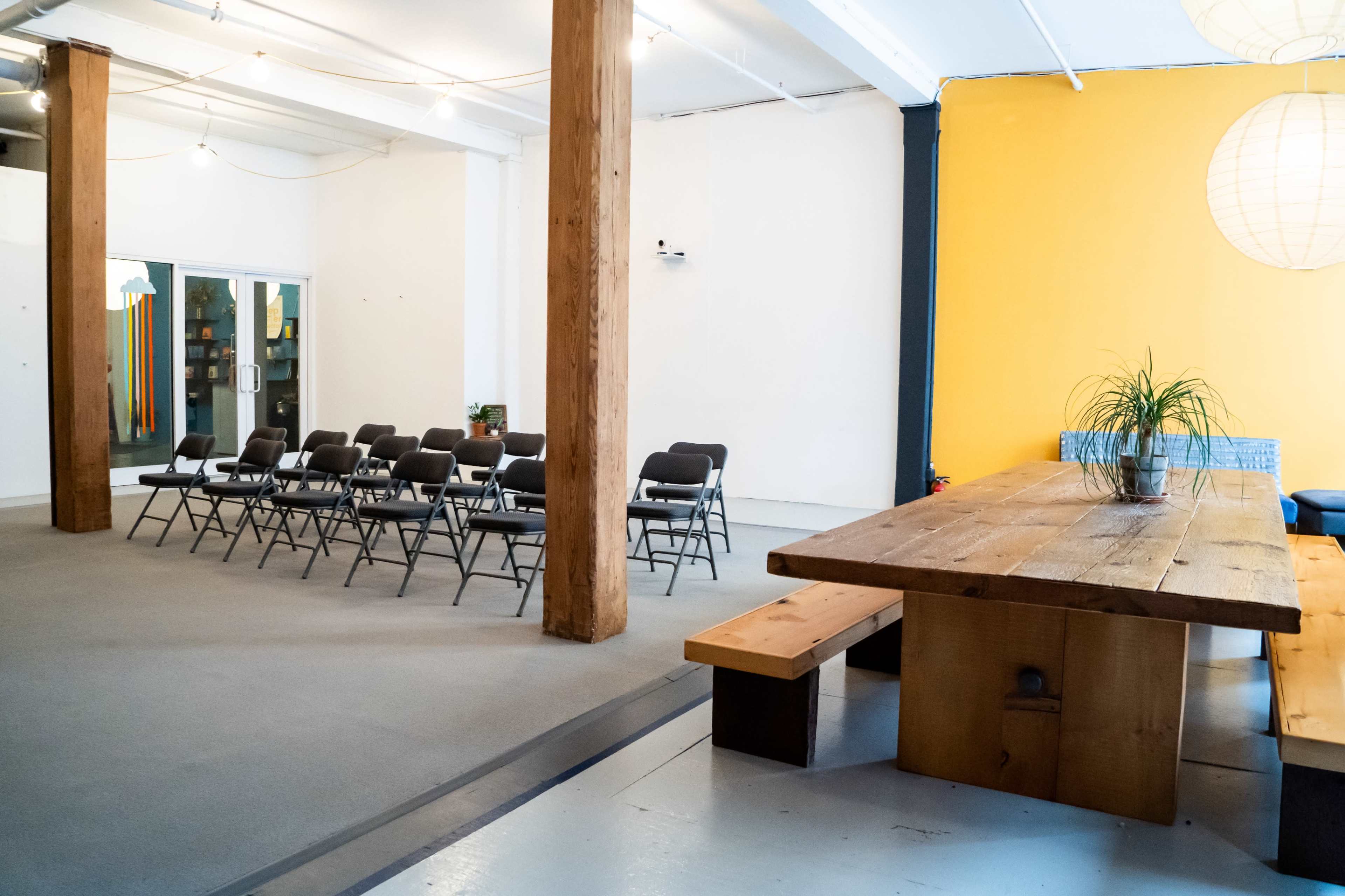 A spacious room with a row of folding chairs facing a large wooden table, featuring an orange accent wall and wooden support pillars.