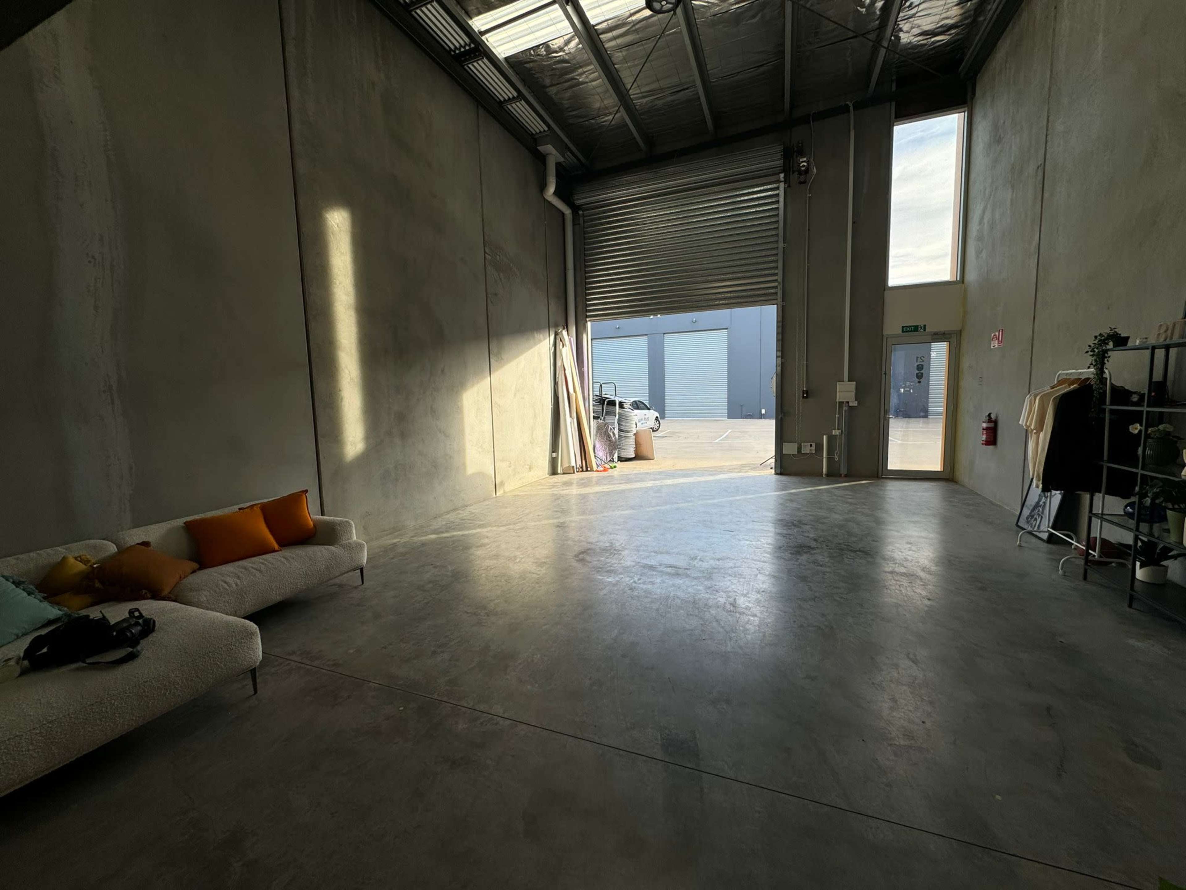 A spacious industrial room features a gray concrete floor, a large roll-up door providing natural light, and a sofa with orange pillows against one wall.