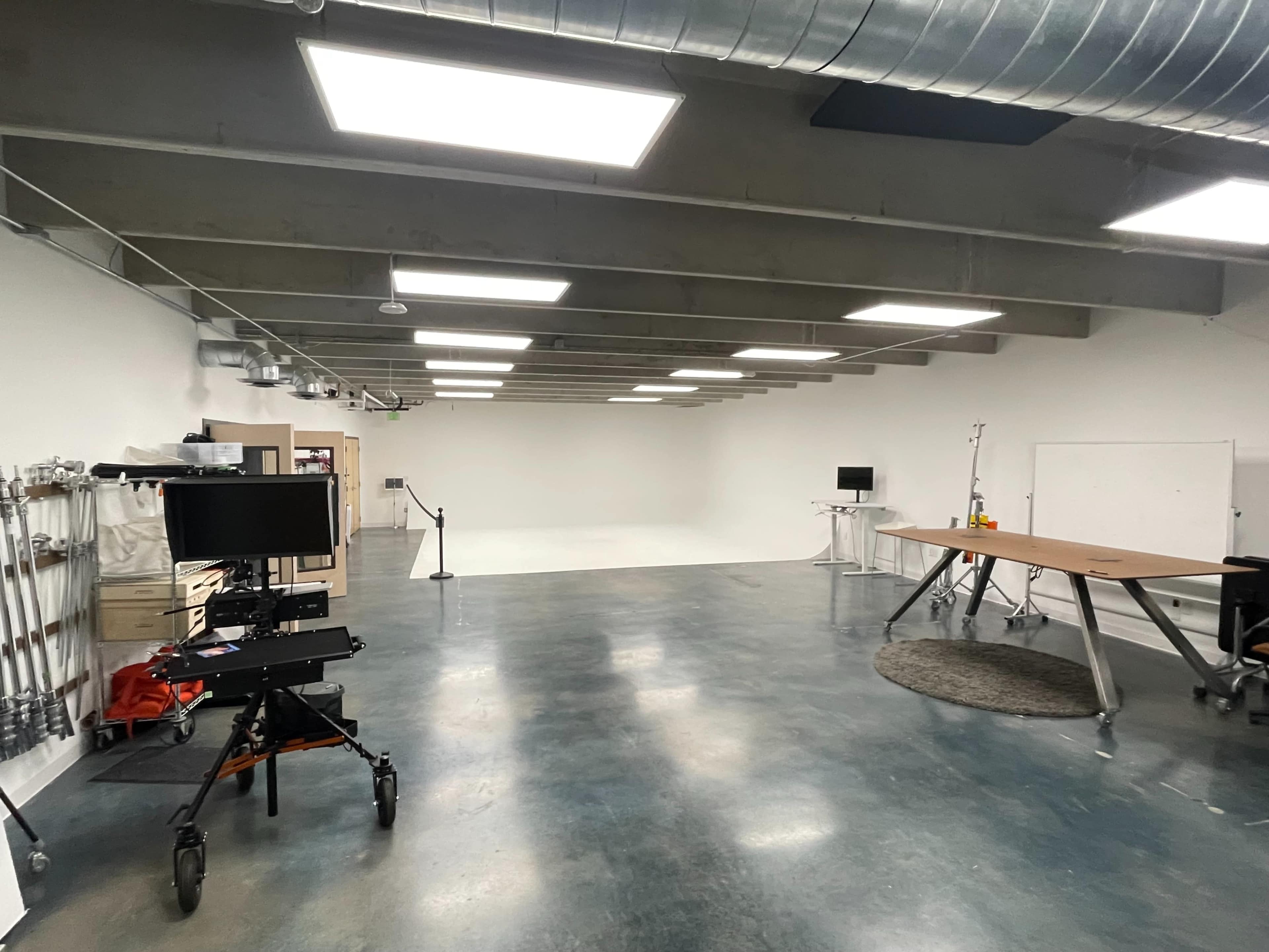 A spacious studio with concrete floors, overhead lighting, and minimal furniture including a large table and equipment on wheels.
