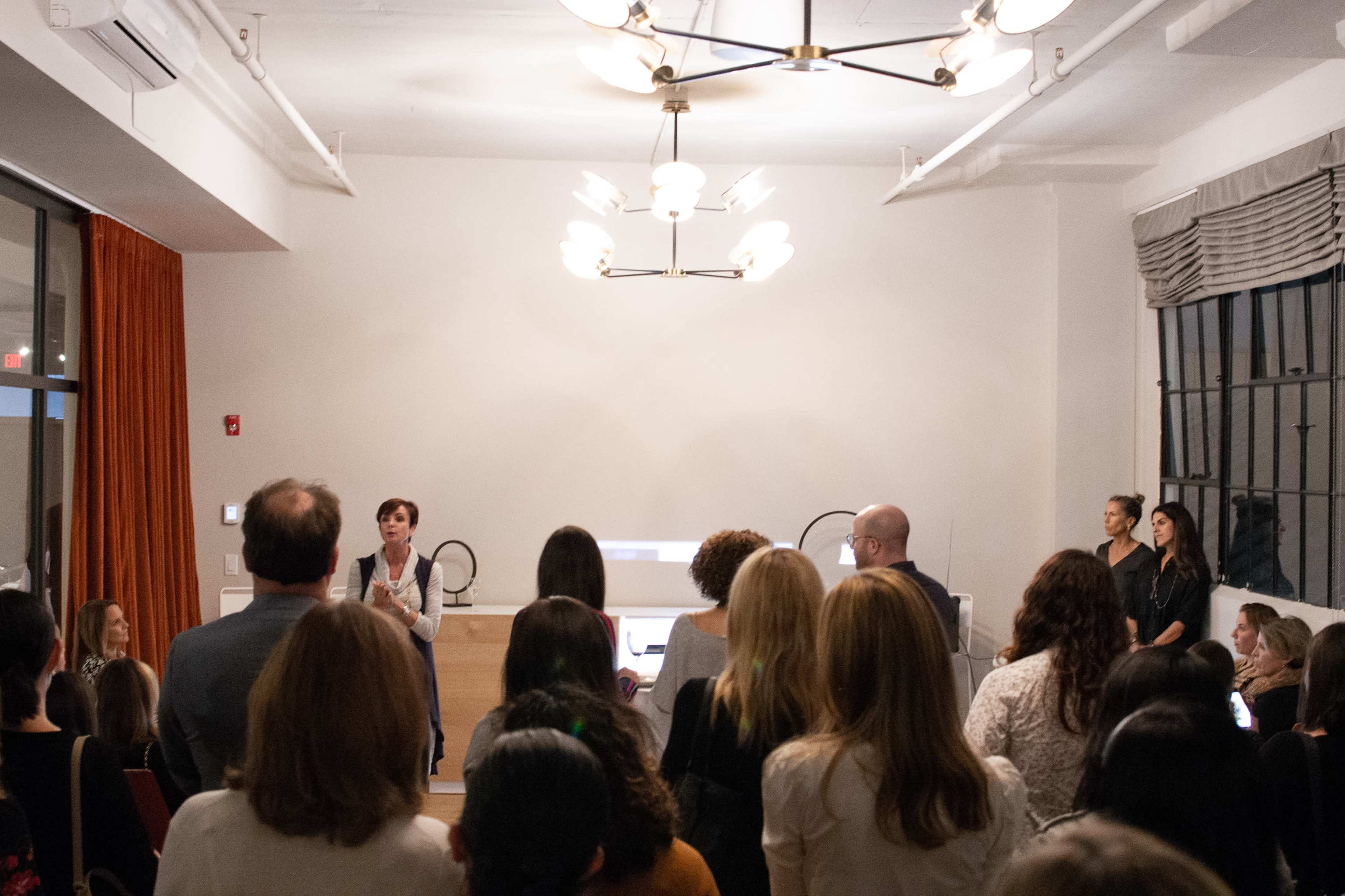 A speaker stands at a wooden podium in front of an audience gathered in a well-lit indoor space.