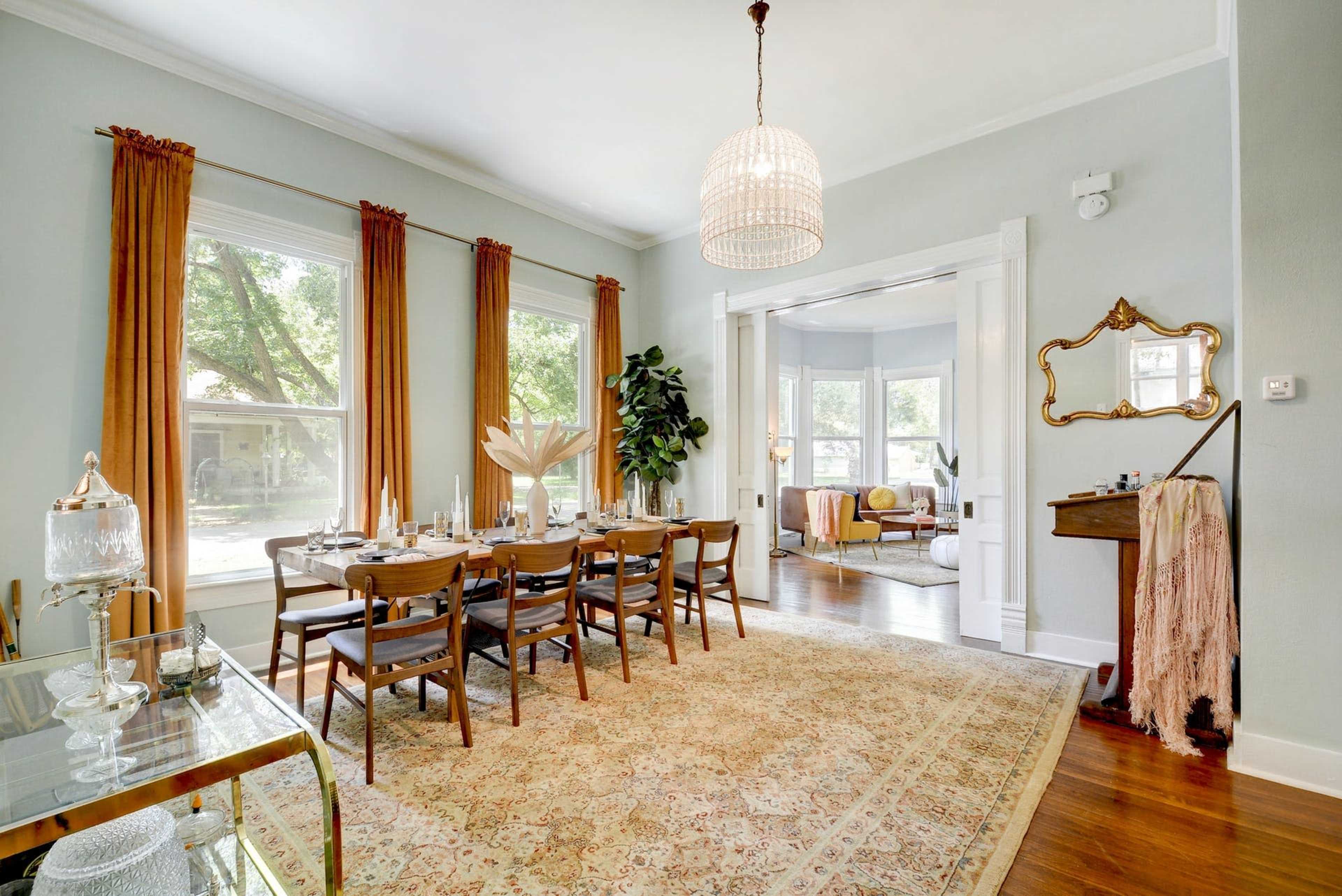A dining room with a large wooden table set for a meal, surrounded by chairs, and featuring large windows with drapes and a chandelier overhead.