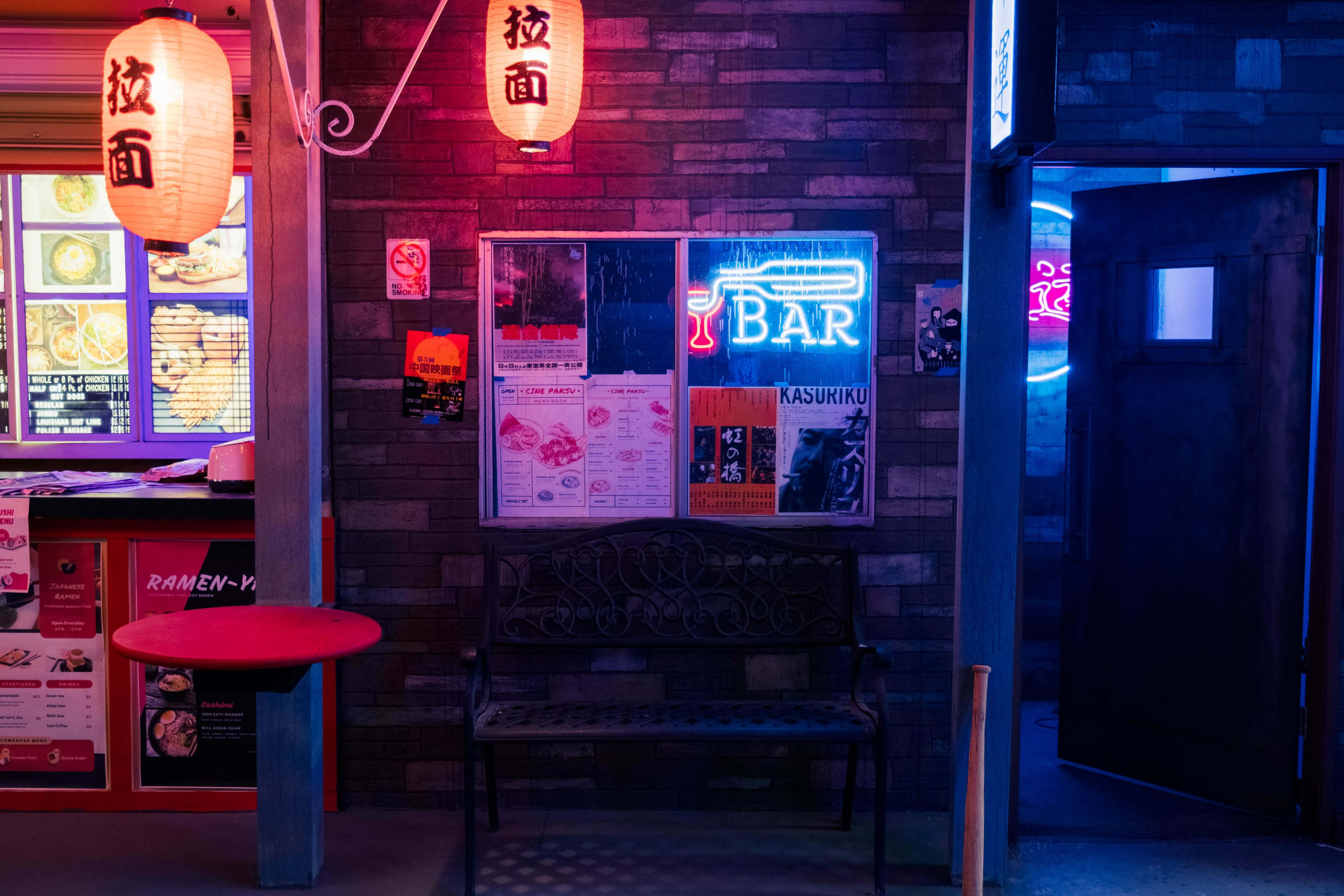 A dimly lit alley features a neon-lit bar sign, colorful lanterns, and a bench beside a wall adorned with various posters.