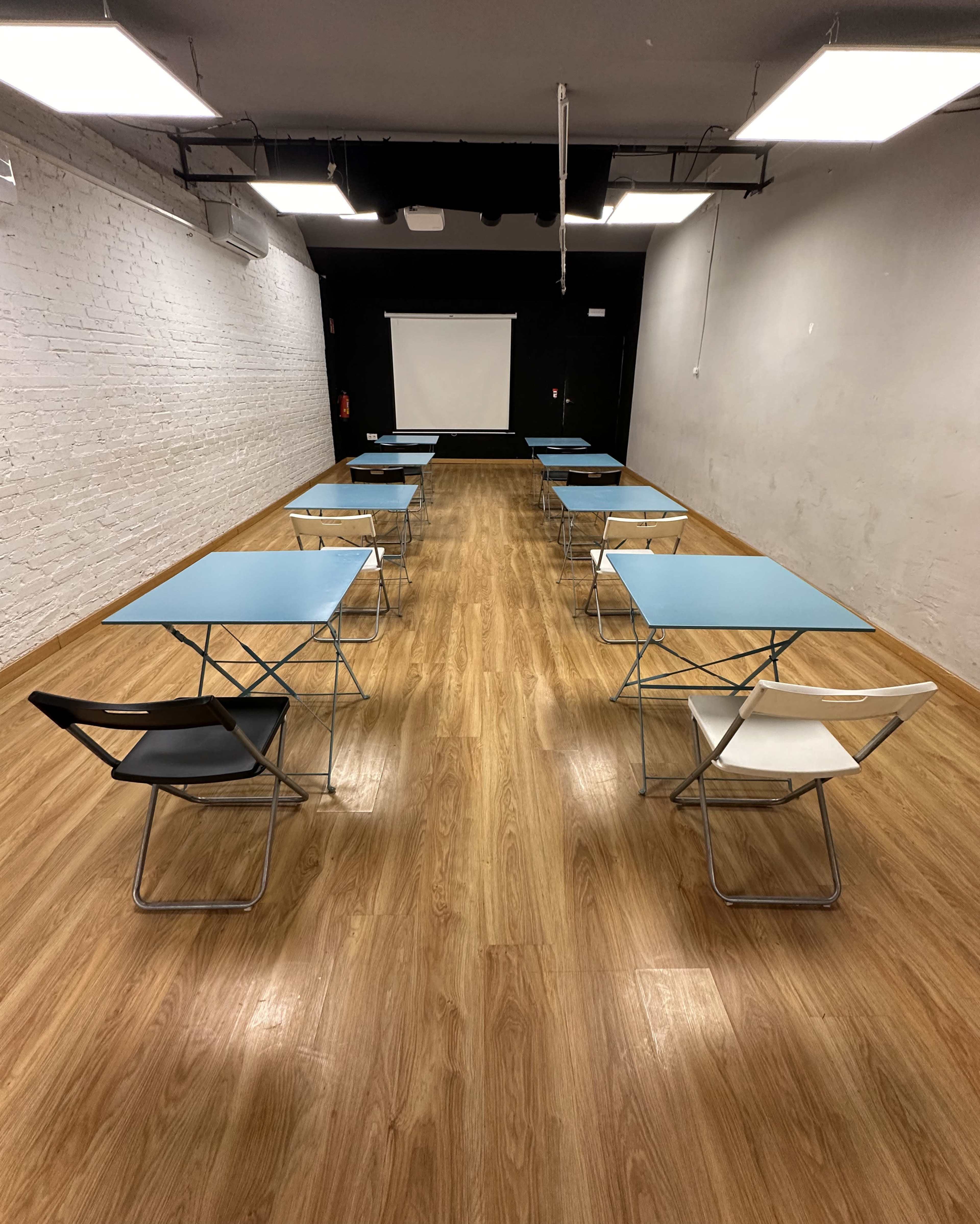 The image shows a row of folding tables with chairs arranged evenly in a spacious, well-lit room featuring a white brick wall and a projector screen at the far end.