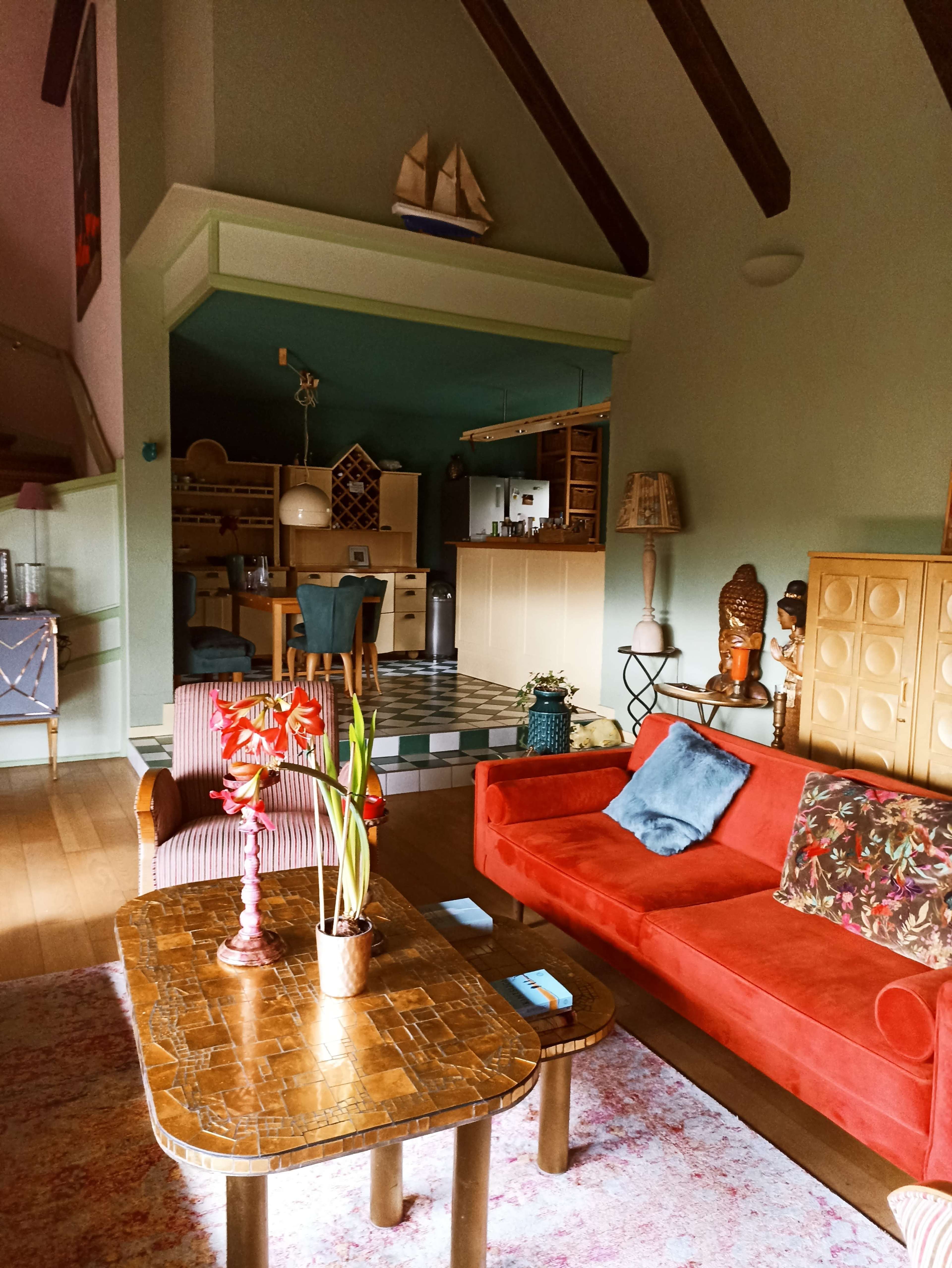 The image shows a cozy living room featuring a red couch, a patterned coffee table, and a kitchen area visible in the background with a checkered floor.