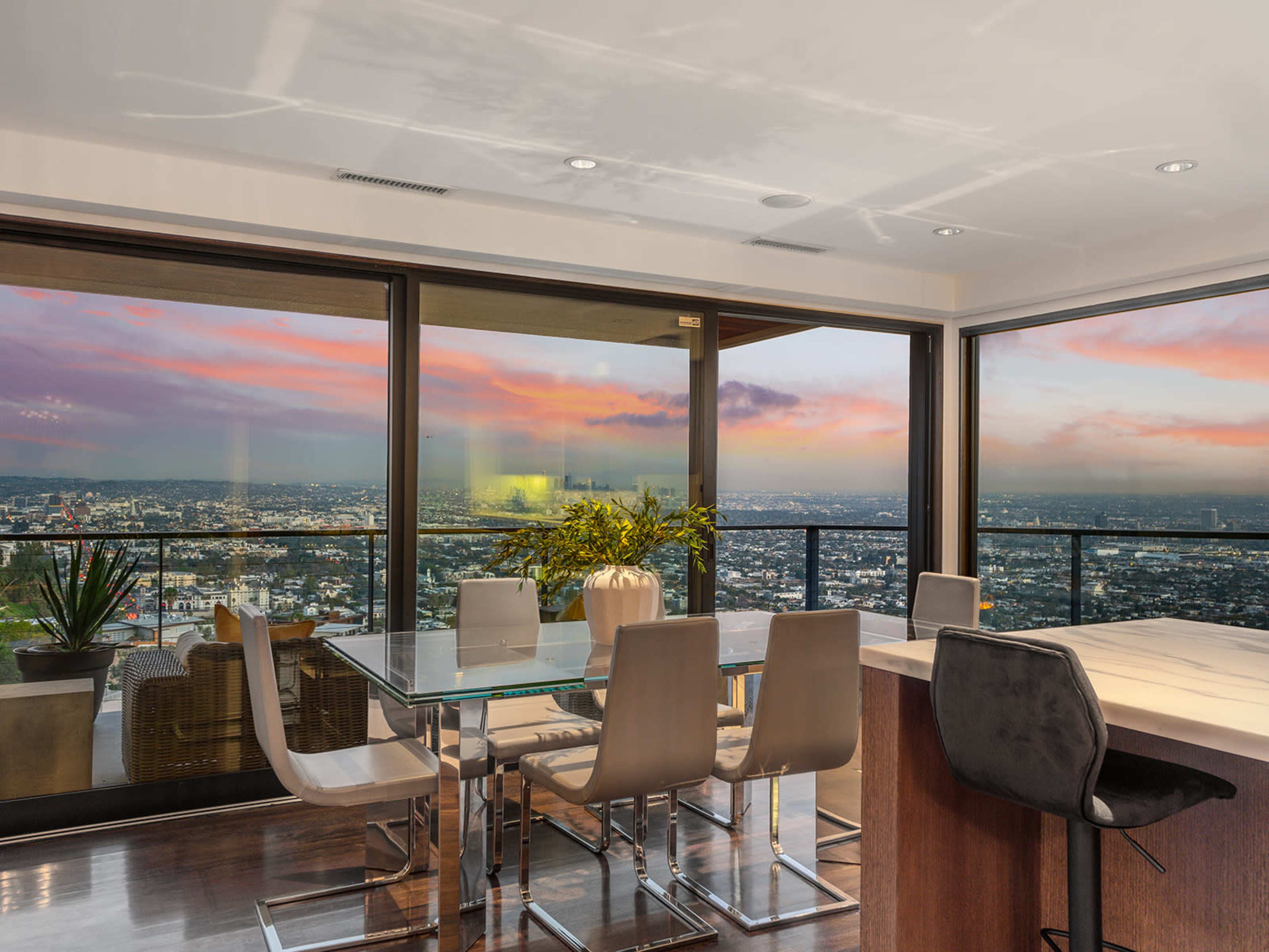 The image shows a modern dining area with a glass table and chairs, overlooking a city skyline at sunset through large windows.